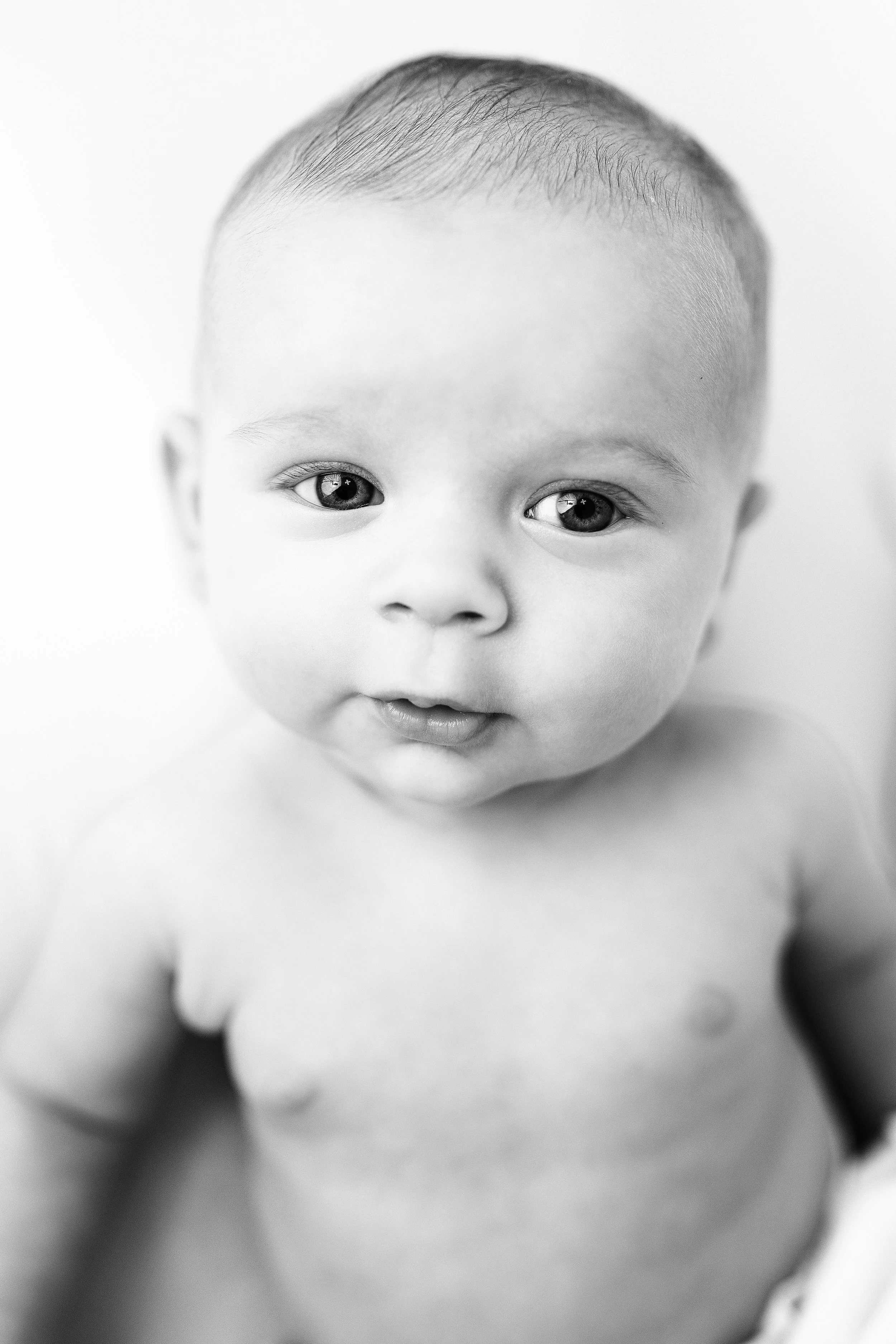 Black and white close-up photograph of an adorable baby with bright eyes and a curious expression, looking directly at the camera.