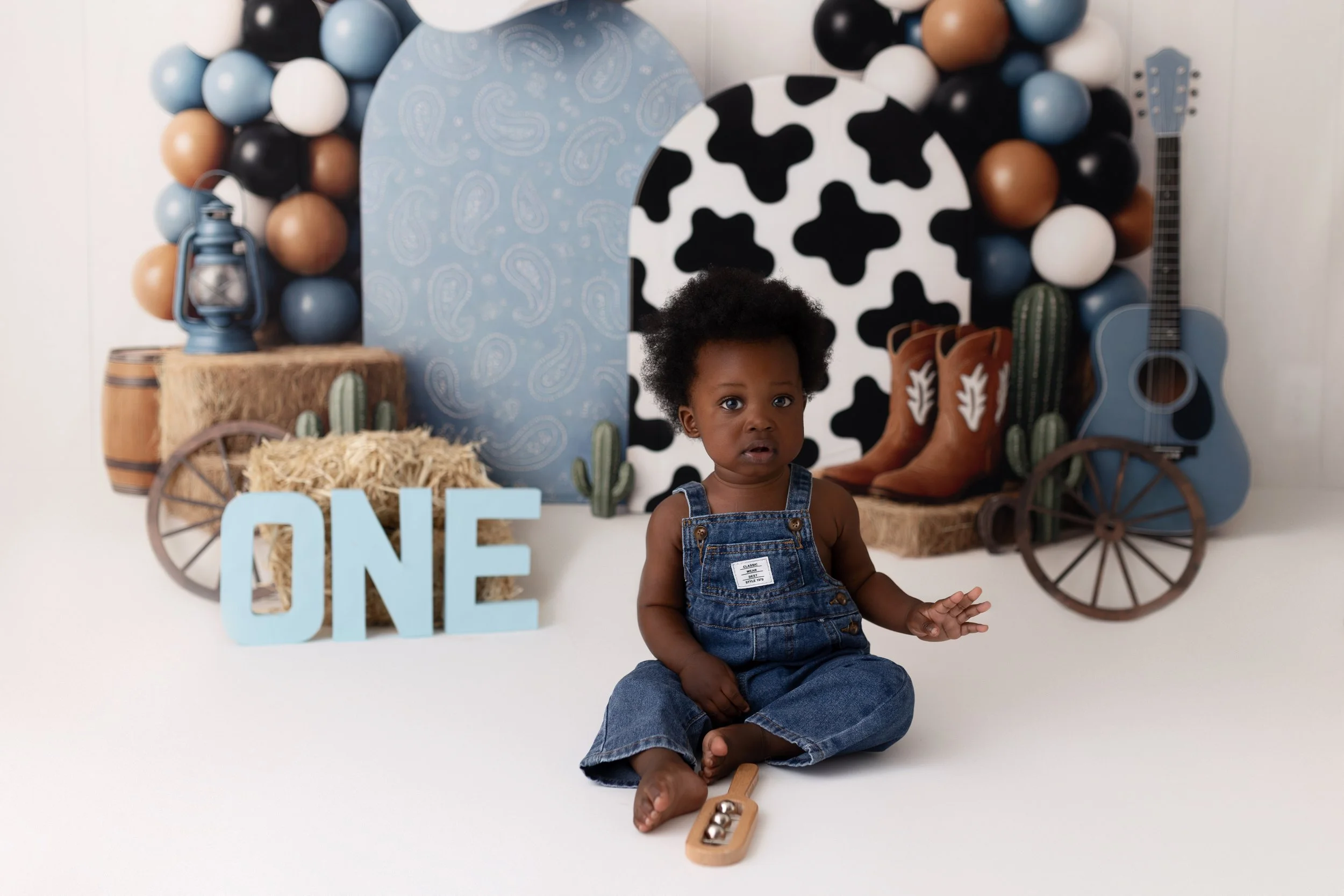 A young child with curly hair, sitting on a white floor in front of a cow-themed cowboy backdrop decorated with balloons, hay, cacti, a guitar, and cowboy boots, with a light blue "ONE" sign.