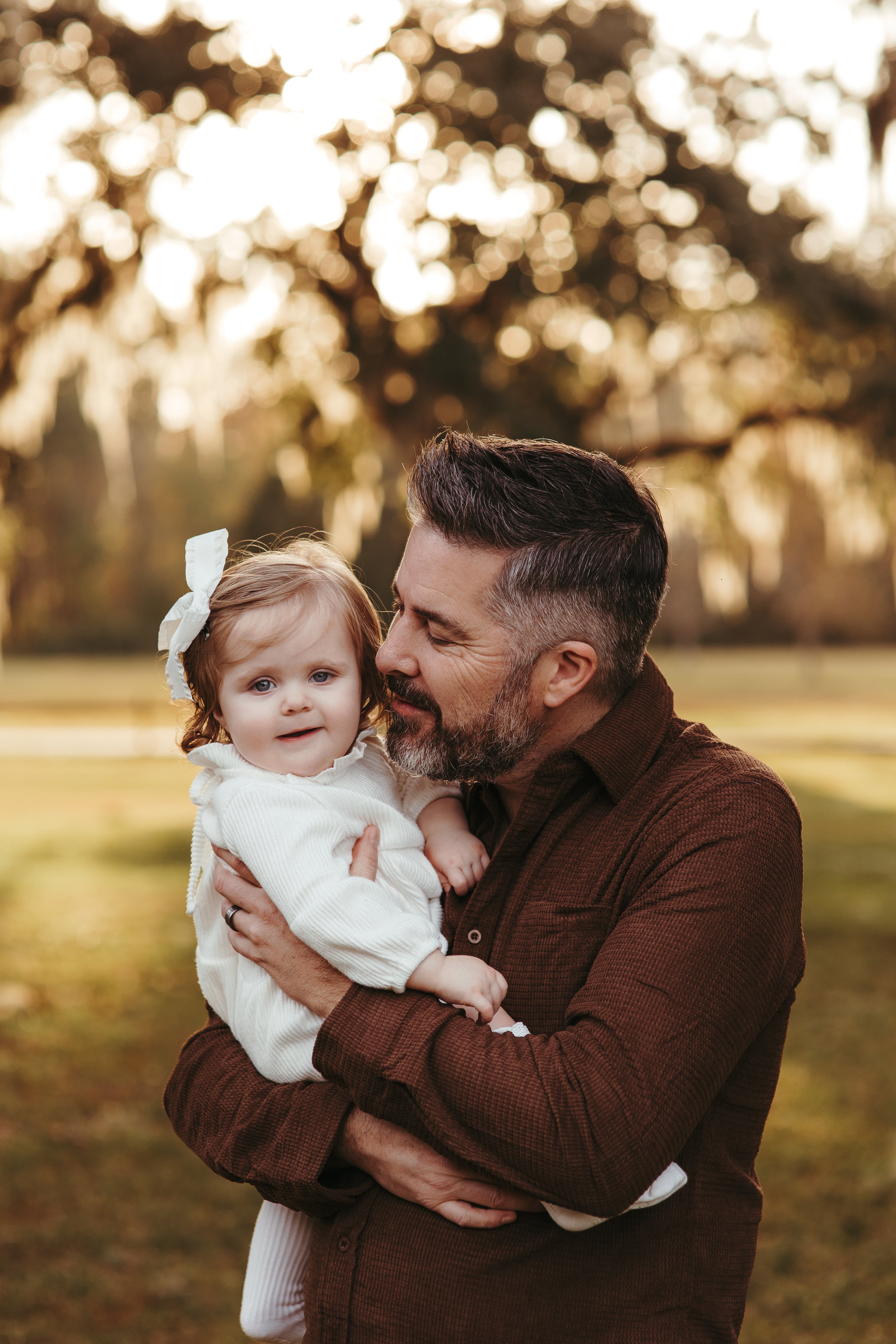 A man with short dark hair and a beard holding a young girl with curly blonde hair and a bow in her hair, outdoors in a park during sunset.