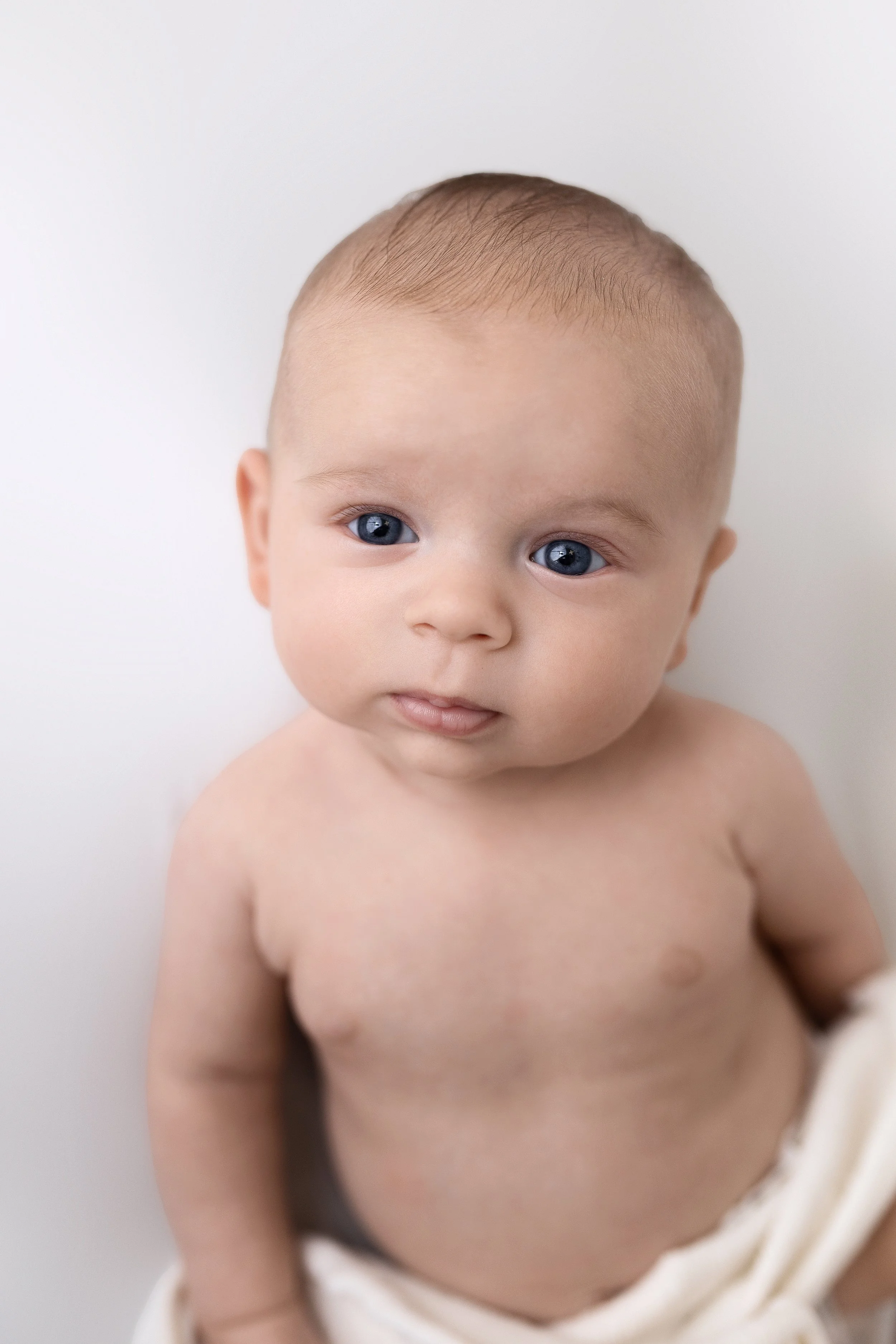Close-up of a young baby with blue eyes, light brown hair, and bare shoulders, looking slightly to the side, against a plain white background.