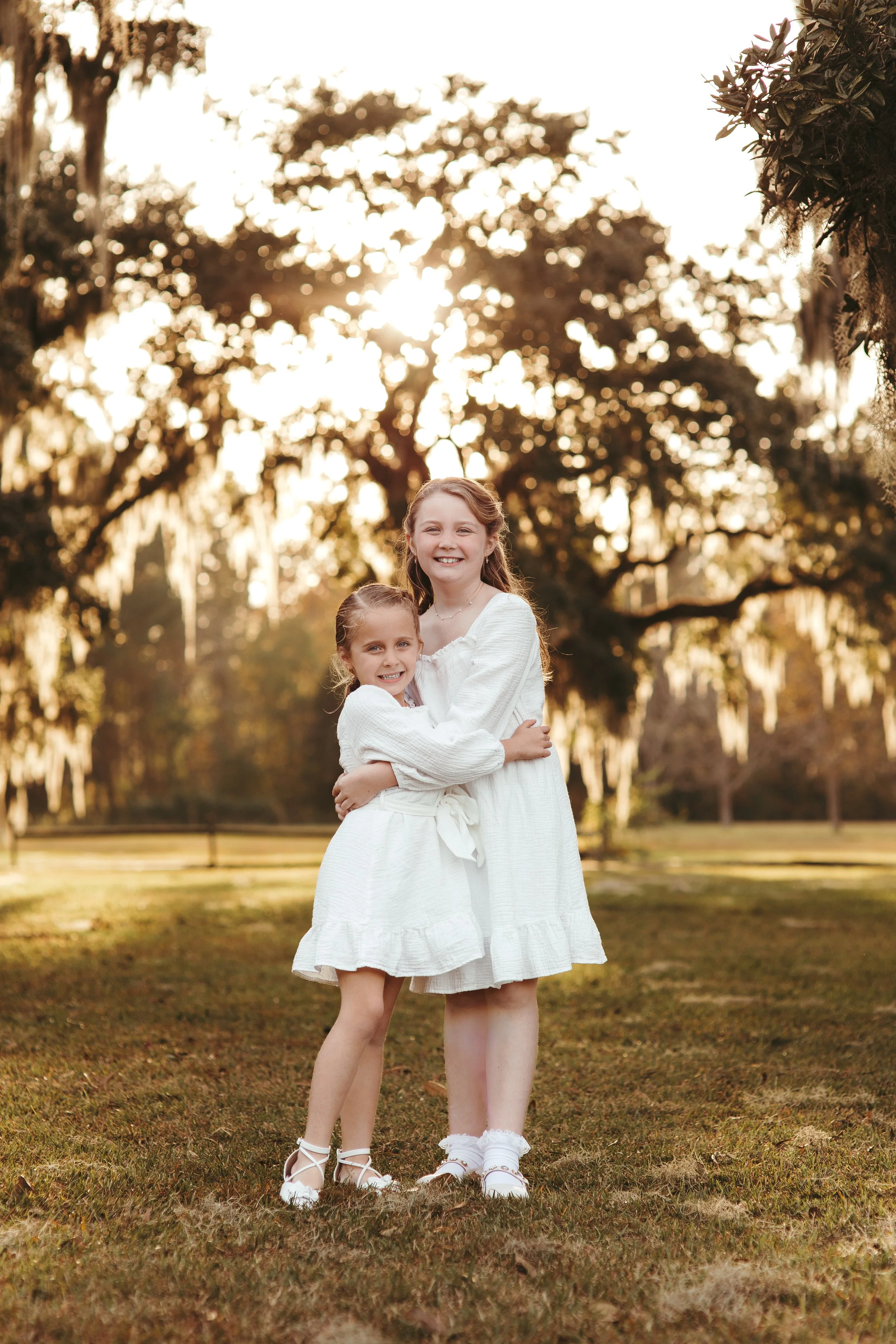 Two young girls dressed in white dresses hugging each other outside during sunset in a park with trees.
