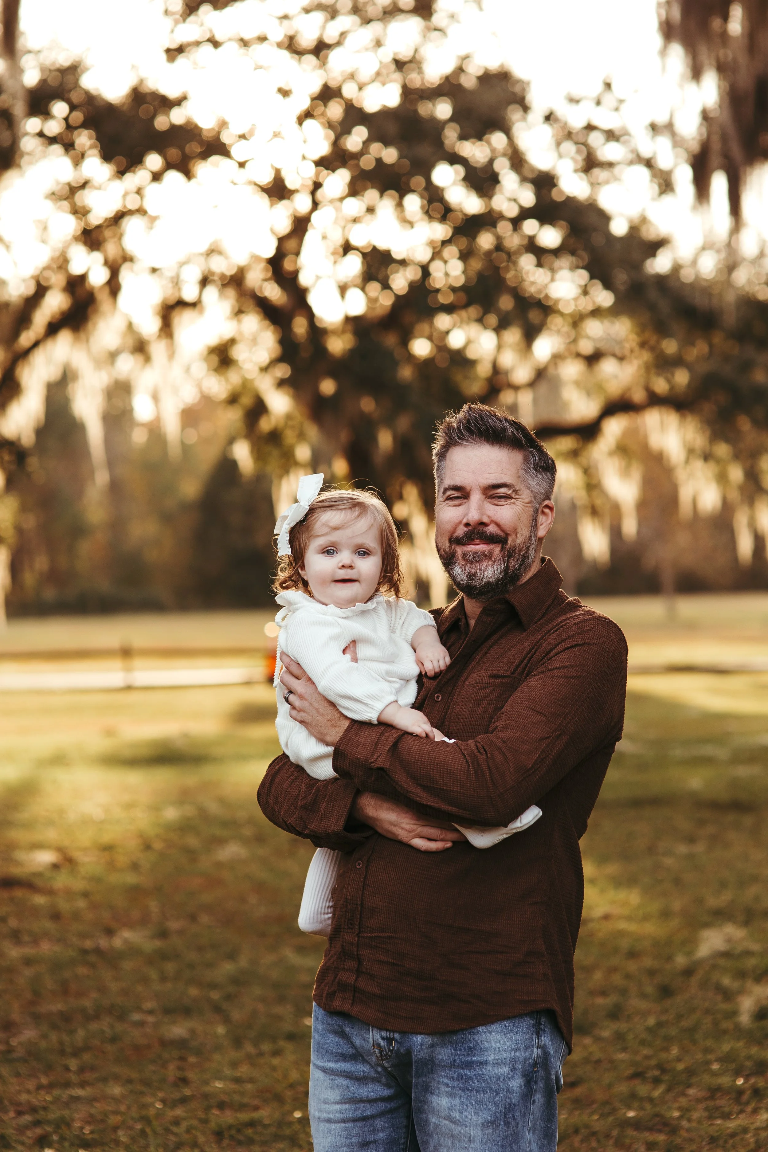 A man holding a young girl outdoors in a park during sunset, with trees and a grassy field in the background.