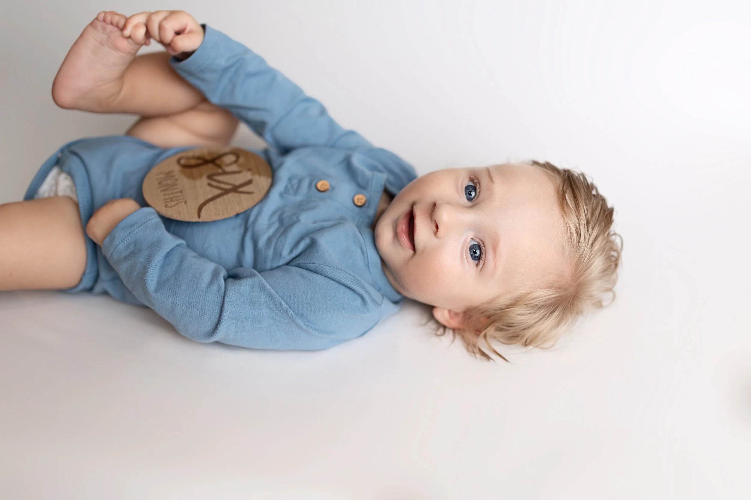 Cute blonde-haired baby boy lying on a white surface, wearing a light blue shirt, with a brown paper tag on his chest with the word 'Love' written on it.