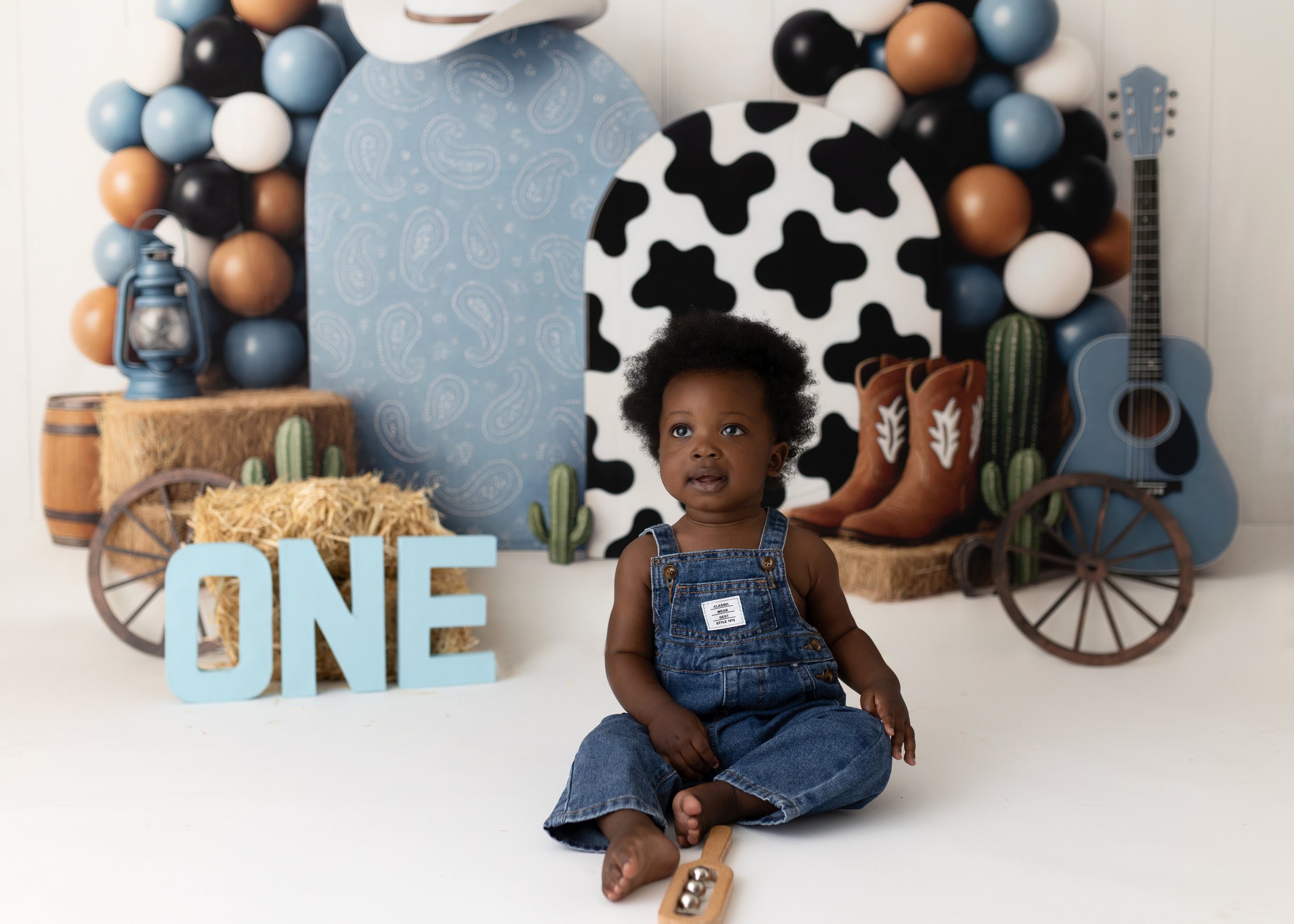 A young child with curly hair sitting on the floor of a country-themed birthday party setup. The background features balloons and large decorative props, including a guitar, cowboy boots, cacti, and a sign spelling 'ONE', indicating a first birthday 
