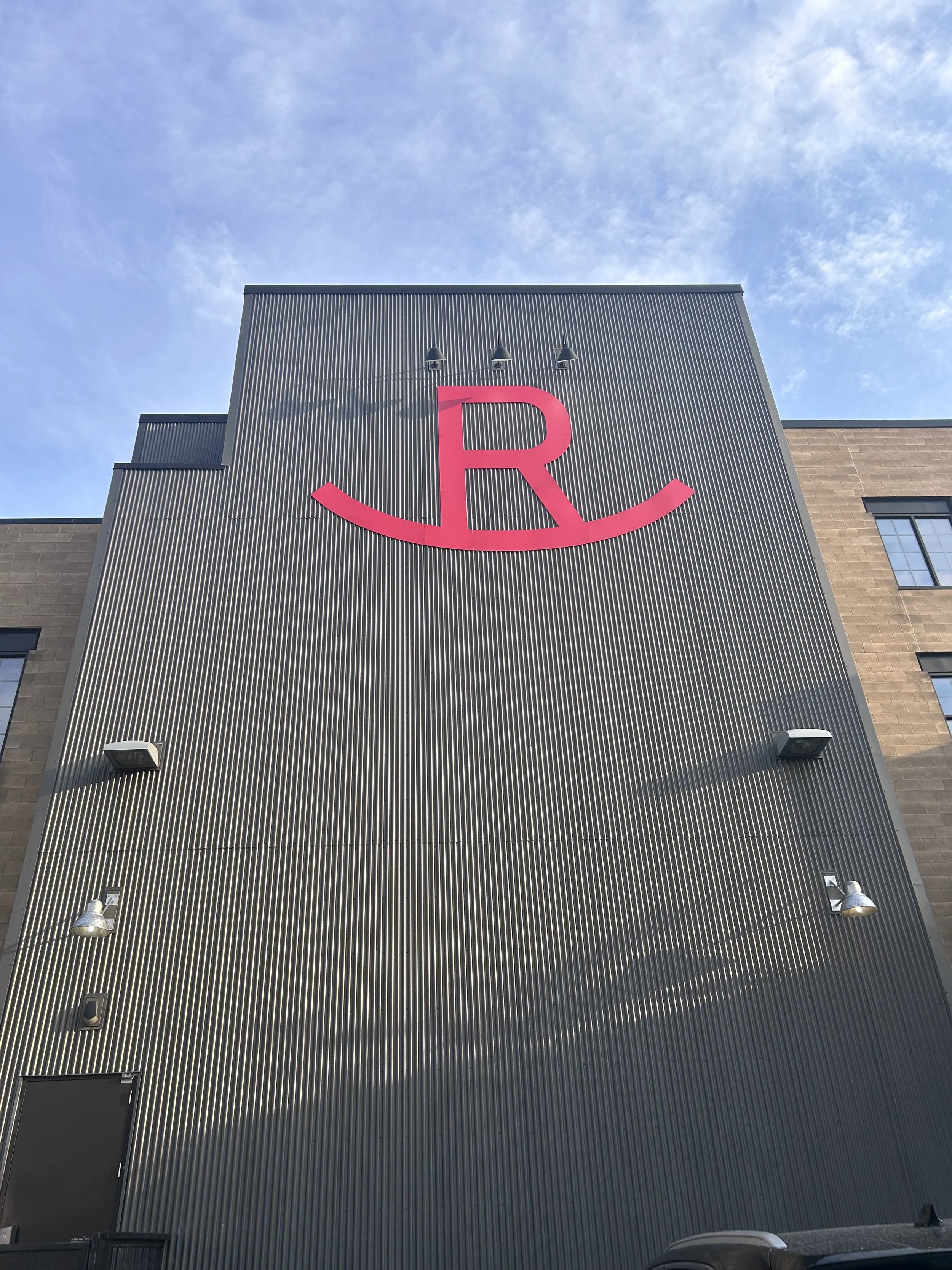 Exterior view of a corrugated metal building facade with a large red letter 'R' and a curved line underneath. The building has security lights and a blue sky is visible.