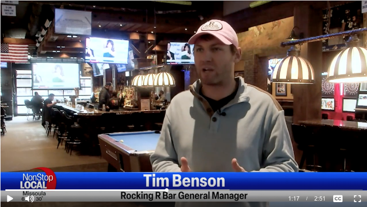 Interior of a bar with a man speaking to the camera. The bar has multiple television screens, pool tables, and is dimly lit. The text at the bottom identifies the person as Tim Benson, General Manager of Rocking R Bar. The "NonStop Local" logo is displayed in the corner with "Missoula" underneath it.