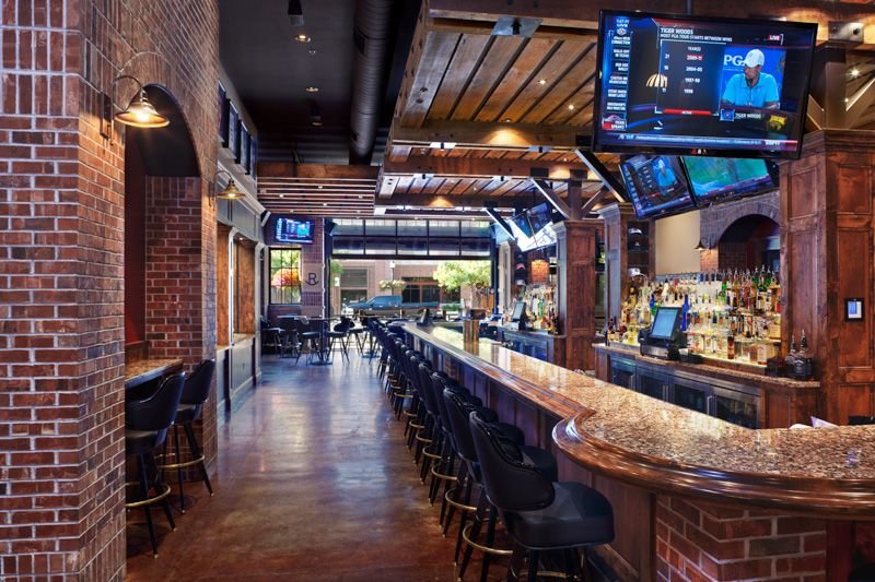 Interior of a stylish bar with brick walls, a long curved bar counter, bar stools, and multiple TV screens displaying sports. The decor features wooden elements and there's a visible entrance with outdoor seating beyond the glass doors.