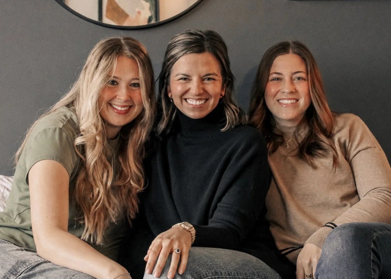 Three women sitting together on a couch, smiling at the camera in a cozy indoor setting with a mirror on the wall behind them.