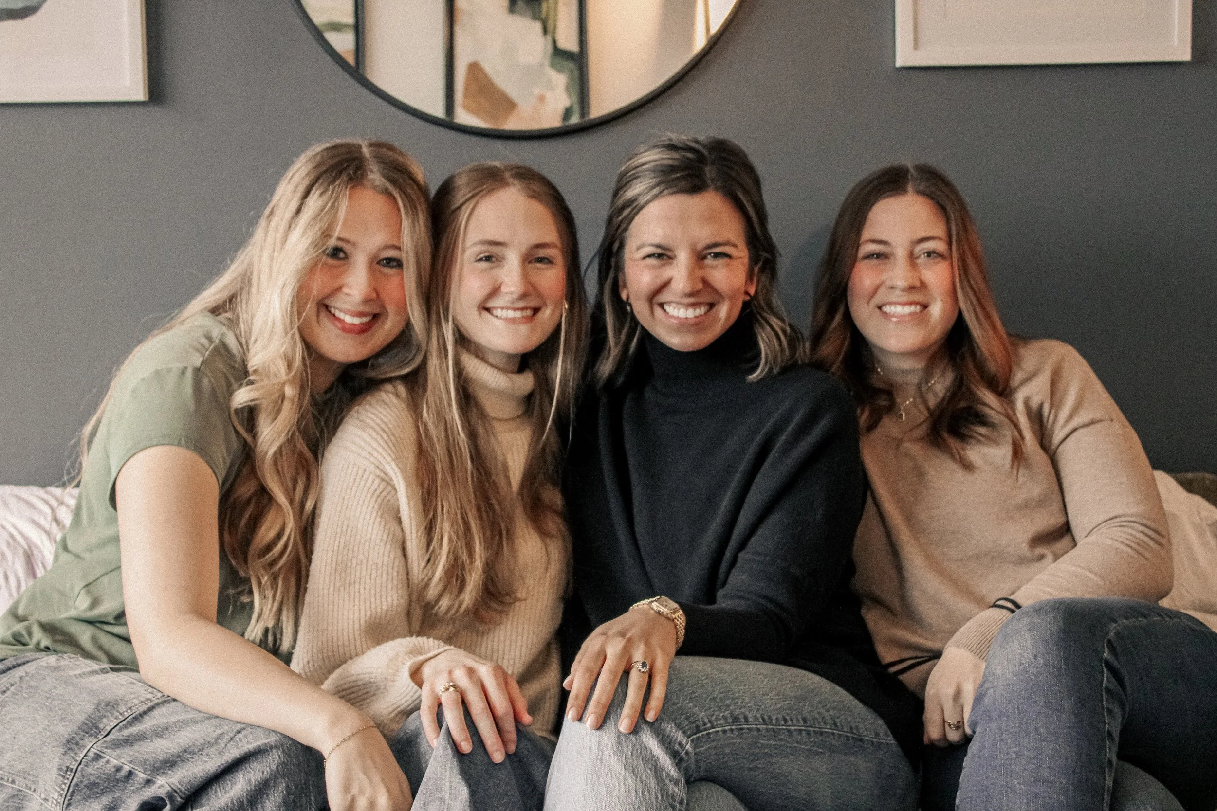 Four women sitting closely together on a couch, smiling at the camera, in a cozy living room with a dark wall and abstract art in the background.