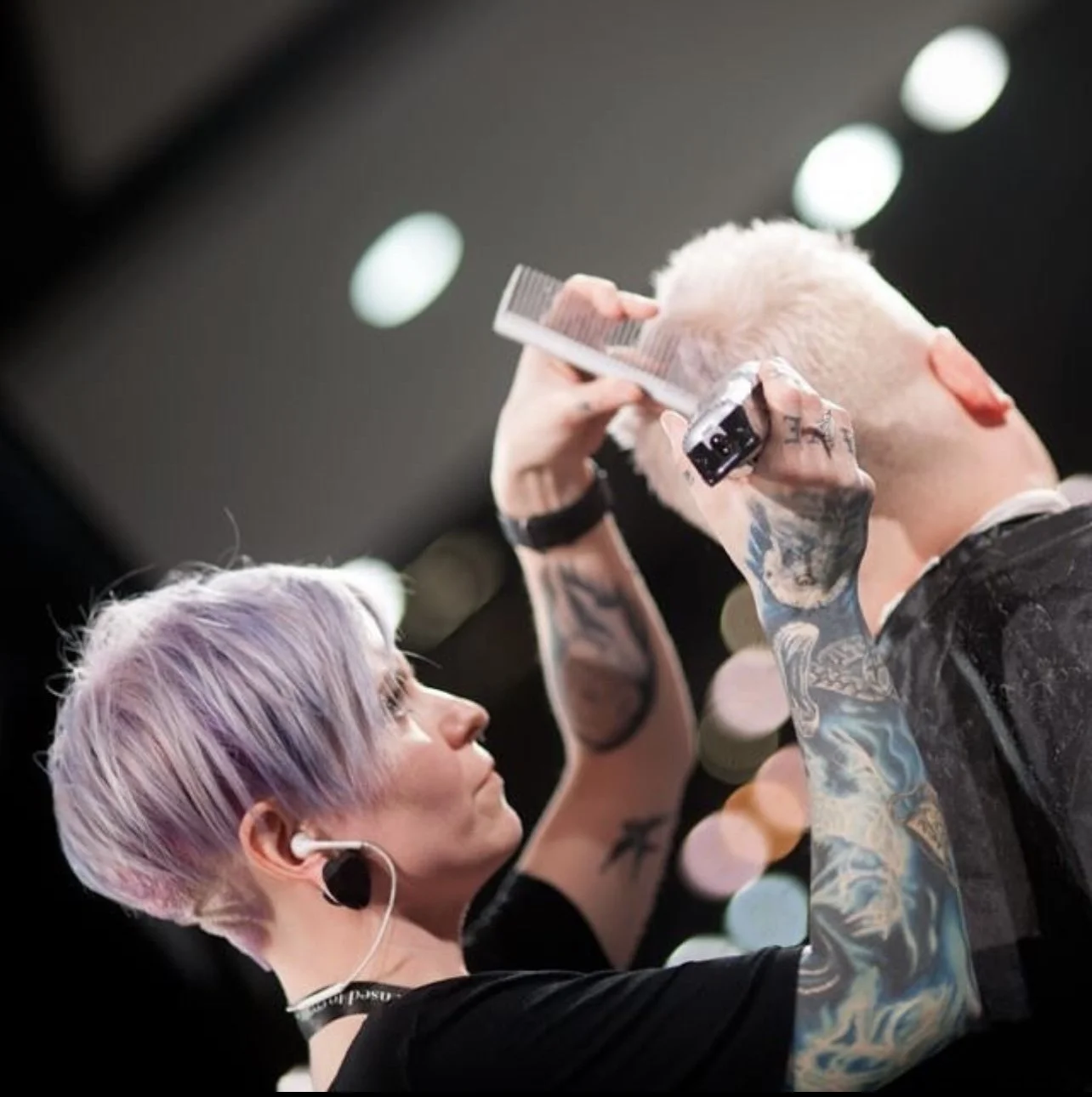 Tattooed hairstylist with lavender hair and earbud creates a textured blond crop haircut using clippers and a comb at Fern&Moody Salon in Chicago.