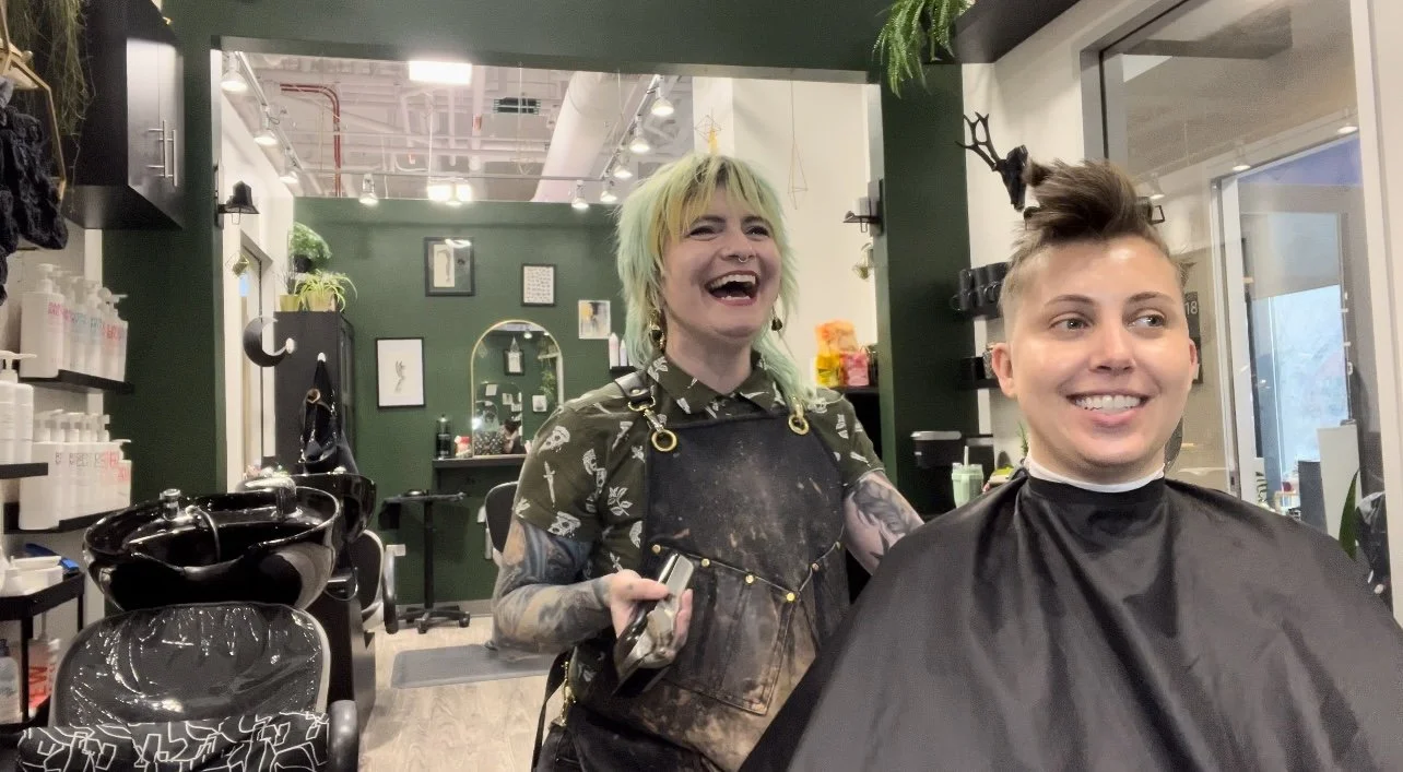 Jackie, a barber at Fern&Moody Salon, laughs with a client mid-haircut in a cozy, plant-filled Chicago salon with dark green walls and tattooed arms.