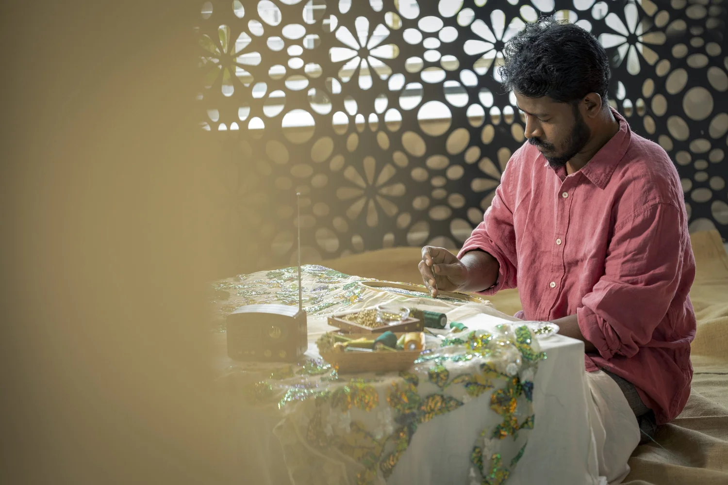 A KUMARI artisan working green and gold botanical embellishment onto white fabric stretched across an embroidery frame.