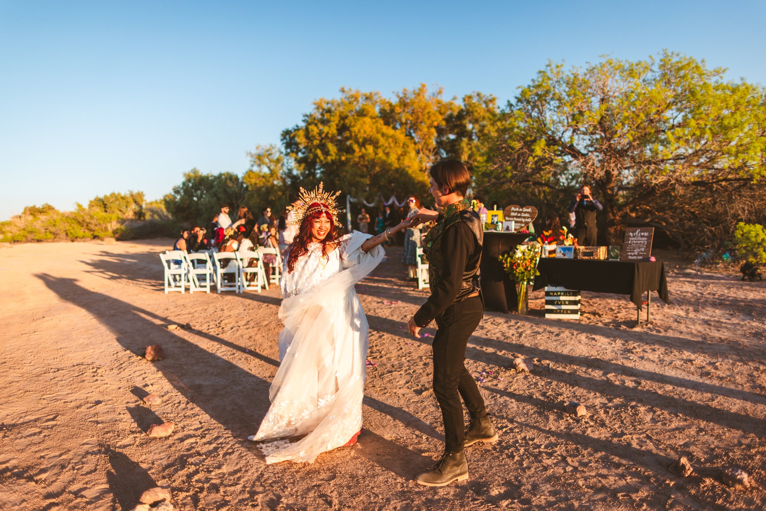A bride and groom dancing outdoors at a wedding ceremony during sunset, with guests seated in the background and a table with wedding signs and decorations nearby.