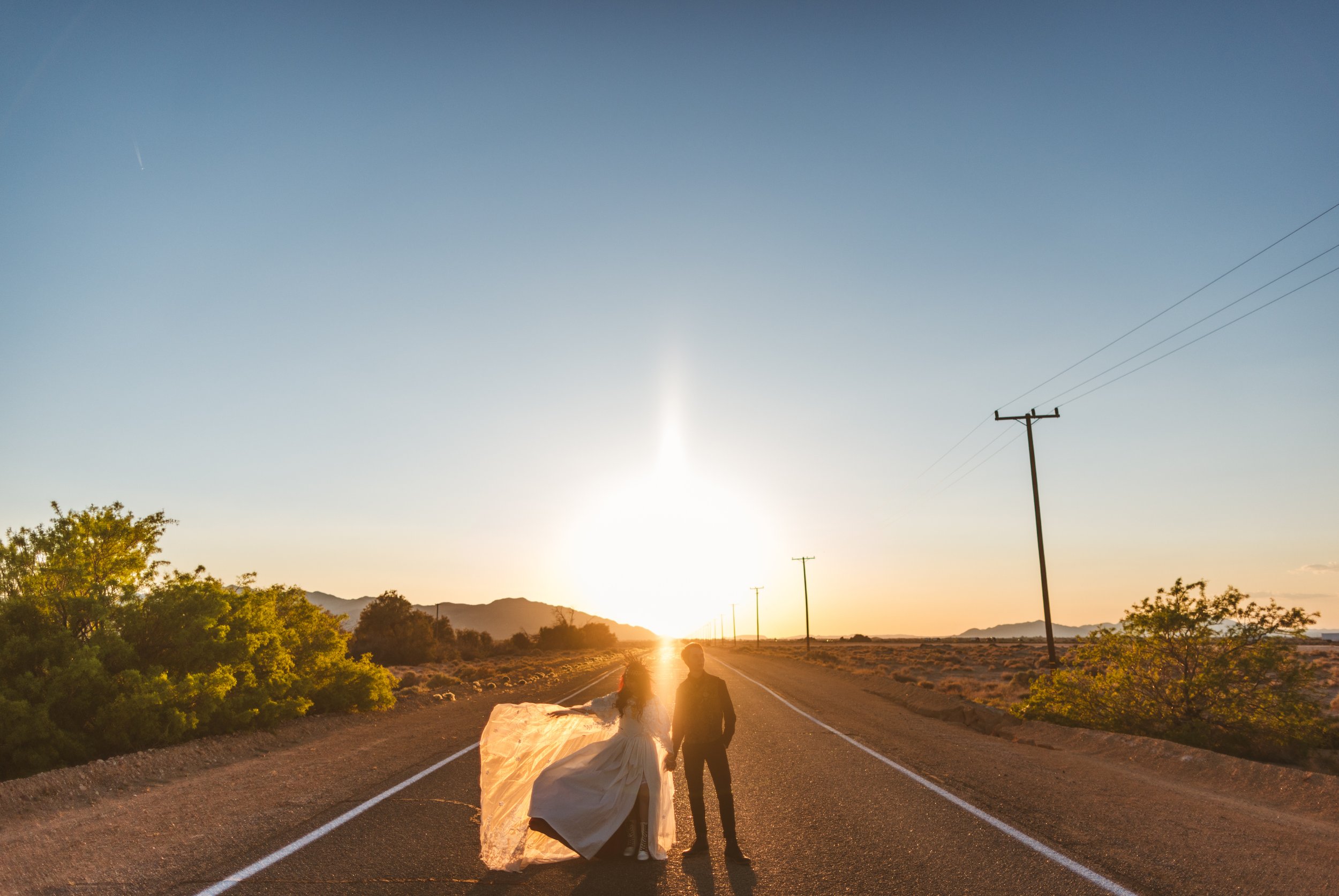 Couple dressed in wedding attire standing on an empty desert road at sunset with mountains and power lines in the background.