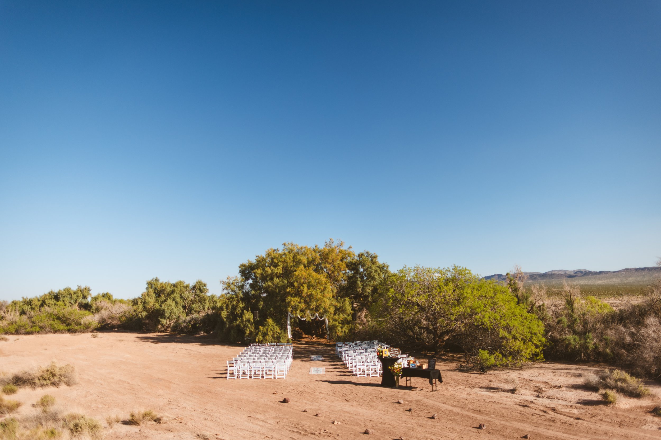 Outdoor desert wedding setup with white chairs arranged in rows facing an arch, under trees, on a dirt ground with mountains in the background.