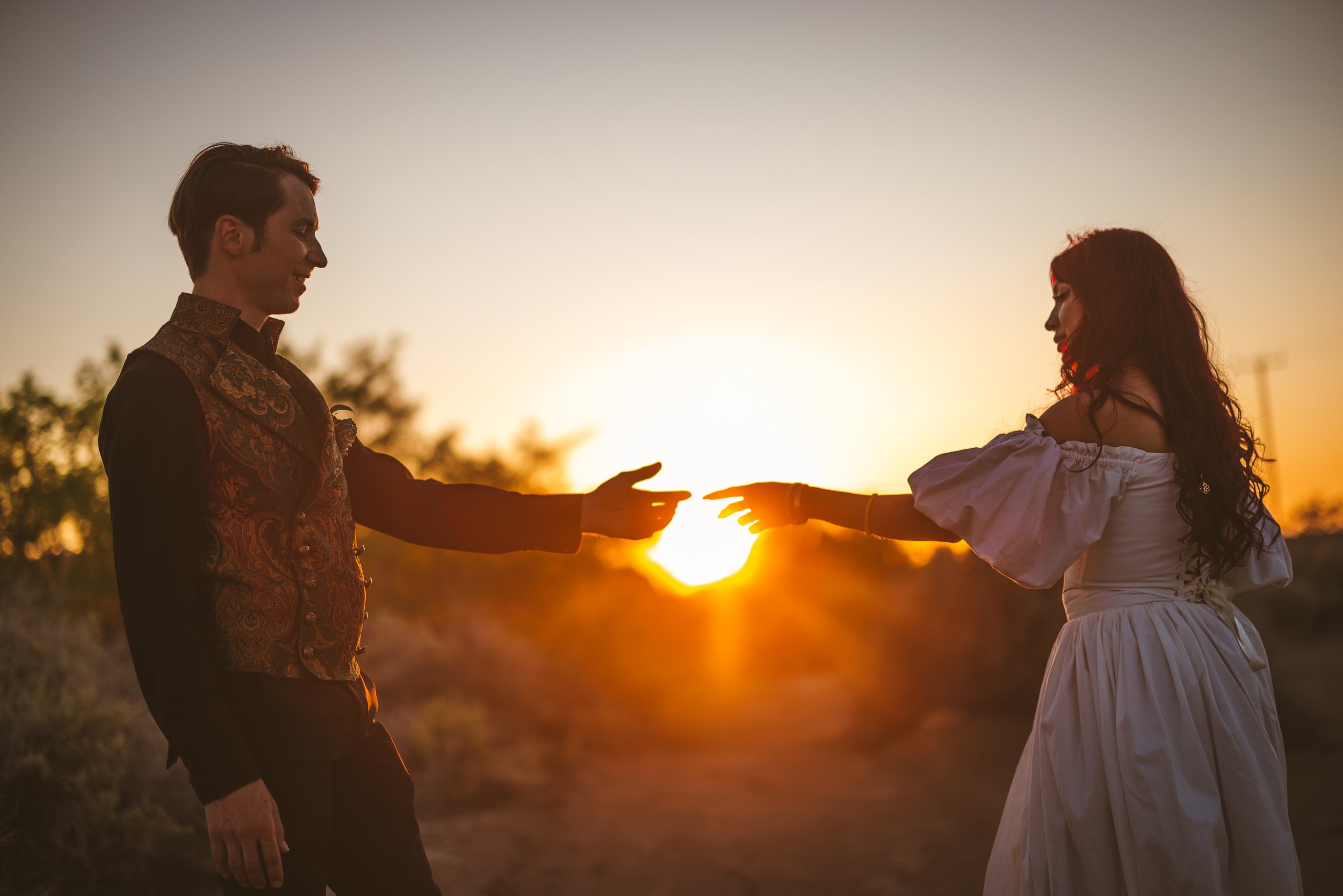 A man and woman reaching towards each other with their fingers touching, standing outdoors during sunset.