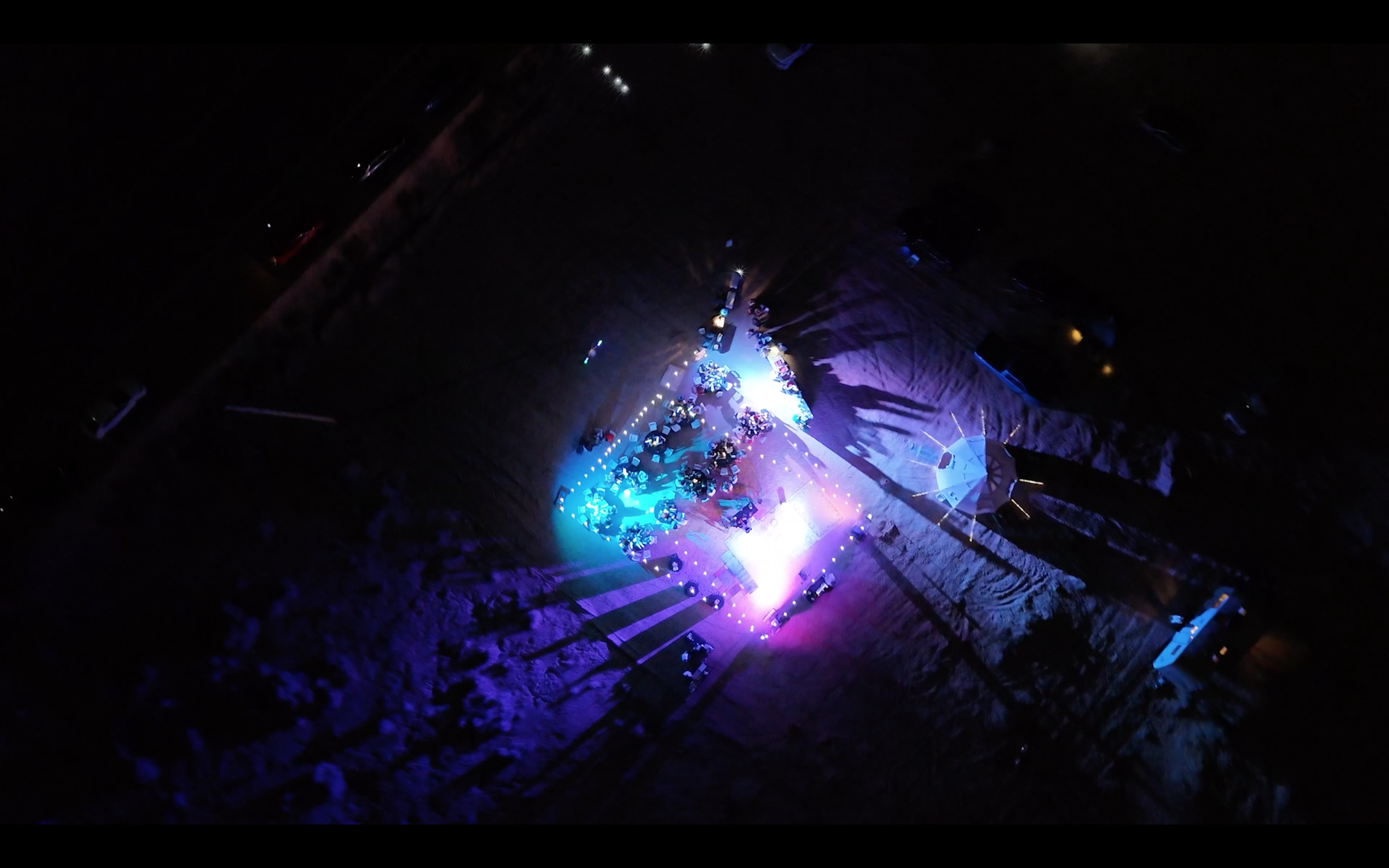 Bird's eye view of an outdoor concert stage illuminated with vibrant purple, blue, and white lights, with musical instruments and performers. The surrounding area appears dark, with the stage lighting creating colorful shadows and reflections on the
