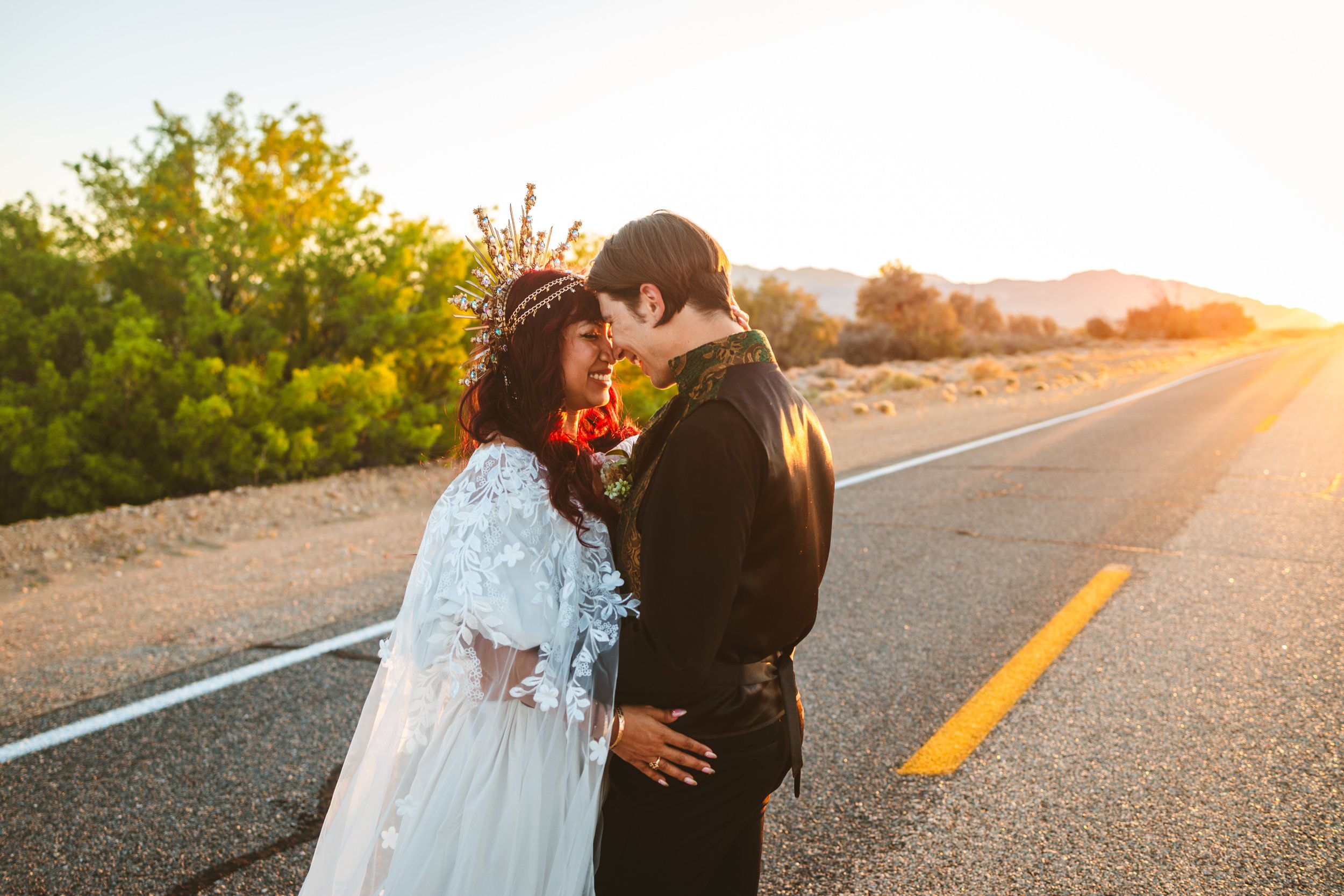 A bride and groom face to face in the middle of an empty road during sunset, smiling and touching foreheads, surrounded by desert landscape and trees.
