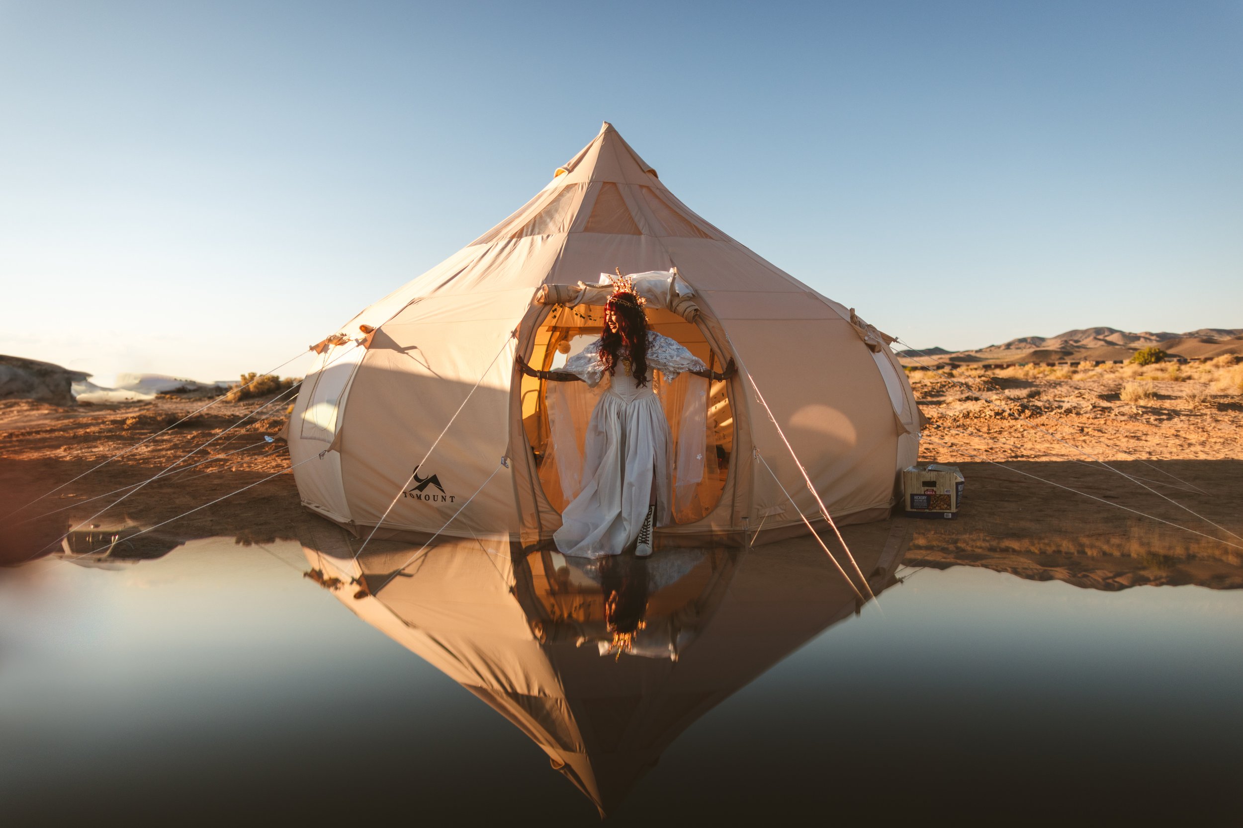 A woman in a white wedding dress and crown standing in the entrance of a beige tent in a desert landscape at sunset, with a reflection of the tent and woman in a water puddle in the foreground.