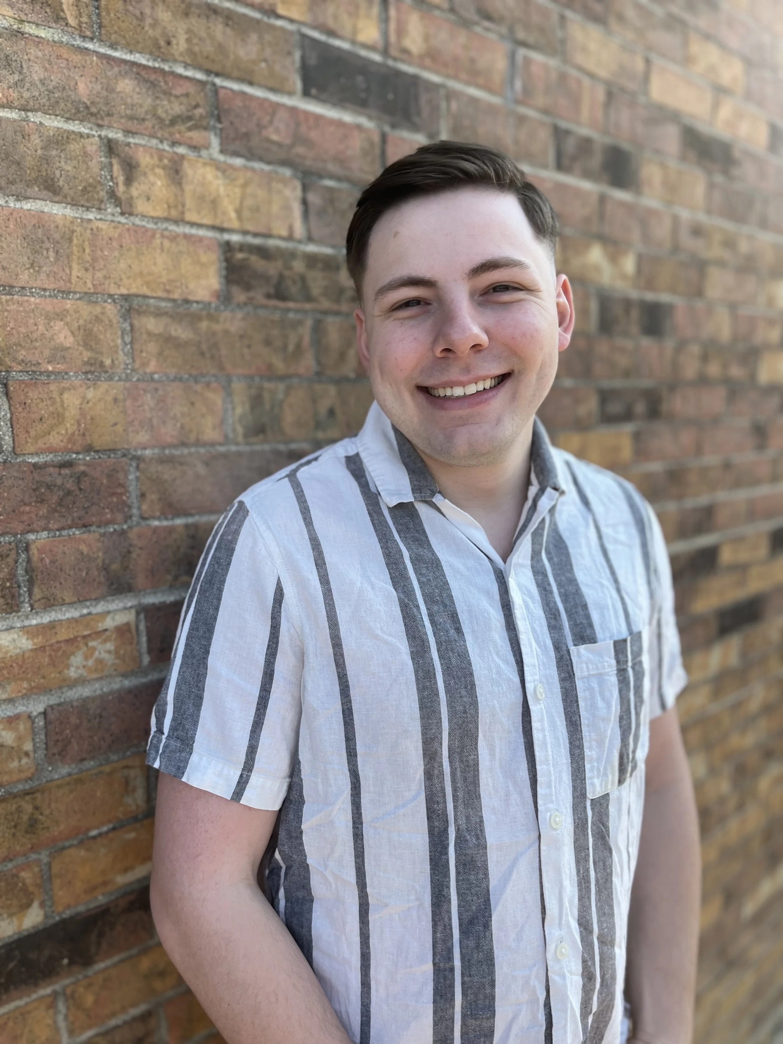A smiling young man wearing a short-sleeved, green and white striped shirt standing against a brick wall.