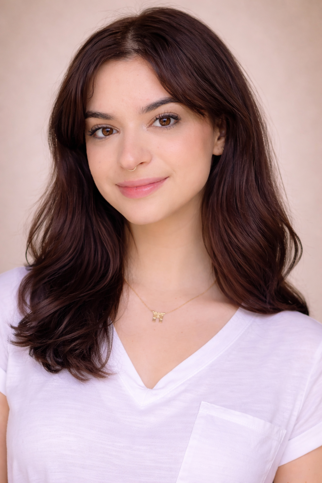 Close-up portrait of a young woman with brown hair and pink highlights, smiling, wearing a black top, earrings, a septum piercing, and a necklace that says 'Strength' against a plain gray background.