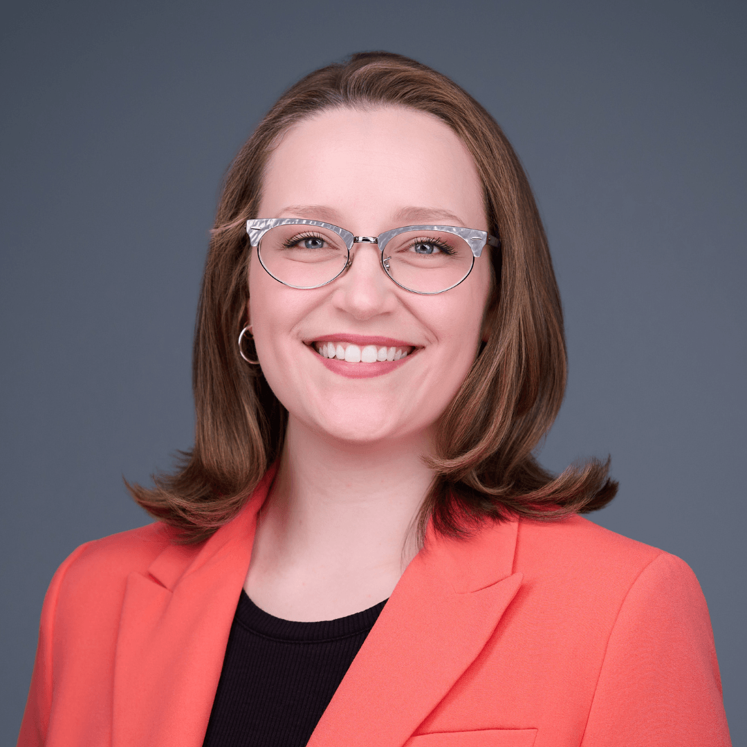 Person wearing glasses and a coral blazer smiling at the camera with a blurred background.