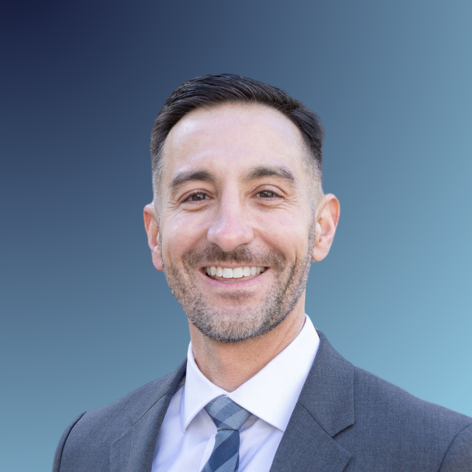 Smiling man in a business suit standing outdoors in front of a stone building.