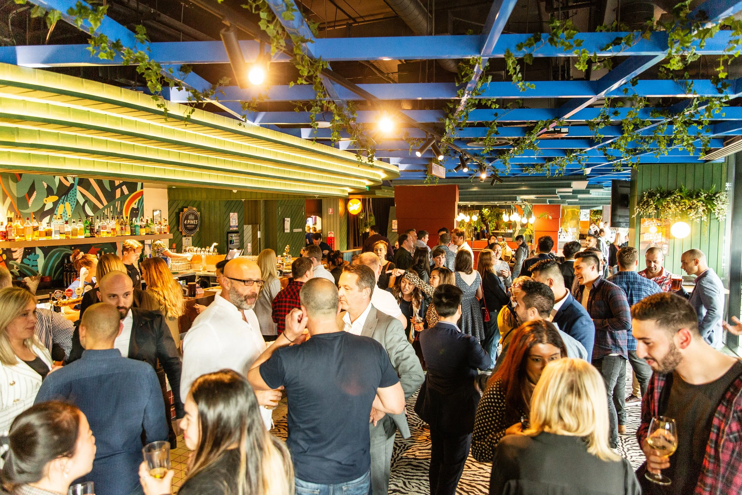 Hightail Bar in the Docklands with people socializing, bar area with various bottles, green and yellow lighting, and ceiling decorated with green plants.