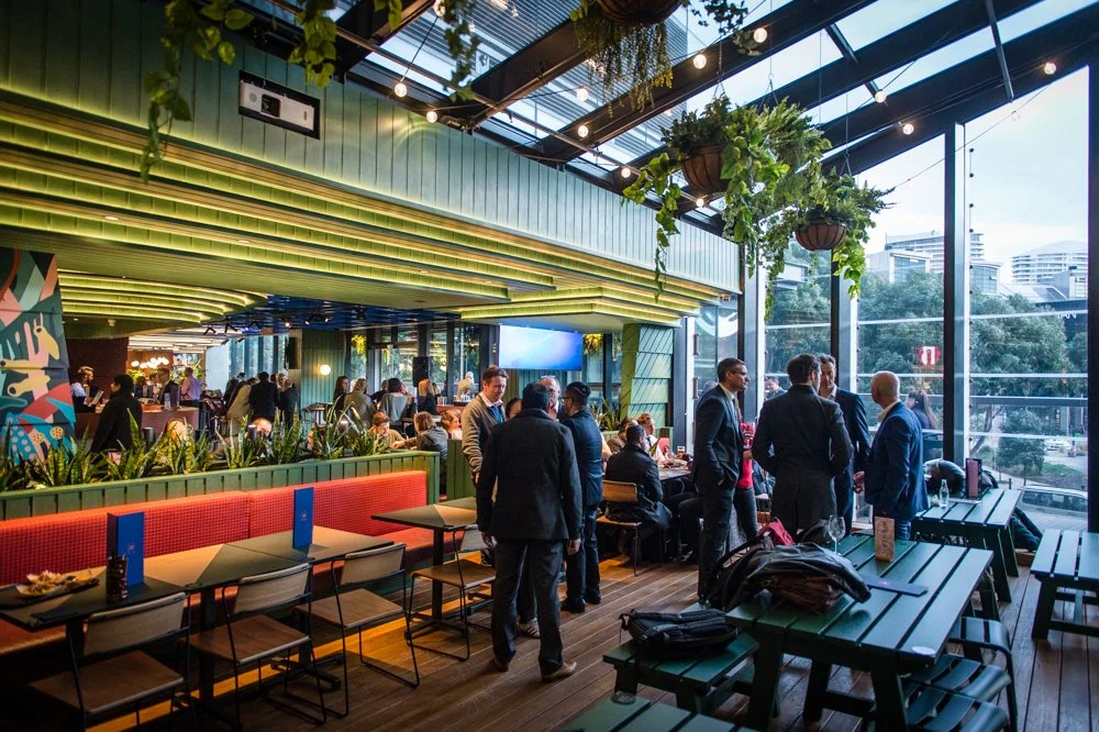 Hightail Bar in the Docklands, corporate function space, with large glass windows, hanging plants, and people in business attire socializing.