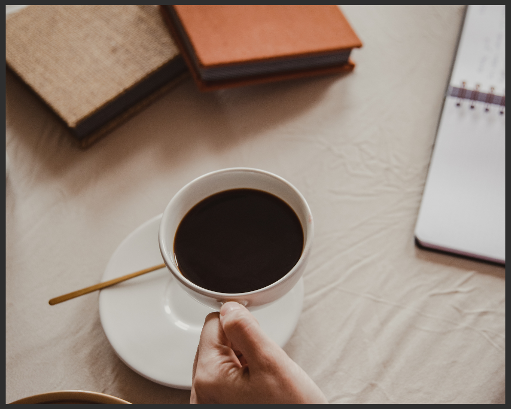 A trauma therapist holding a cup of black coffee on a white saucer with a gold spoon resting on it, on a beige table. In the background, there are closed notebooks and an open notebook or planner.