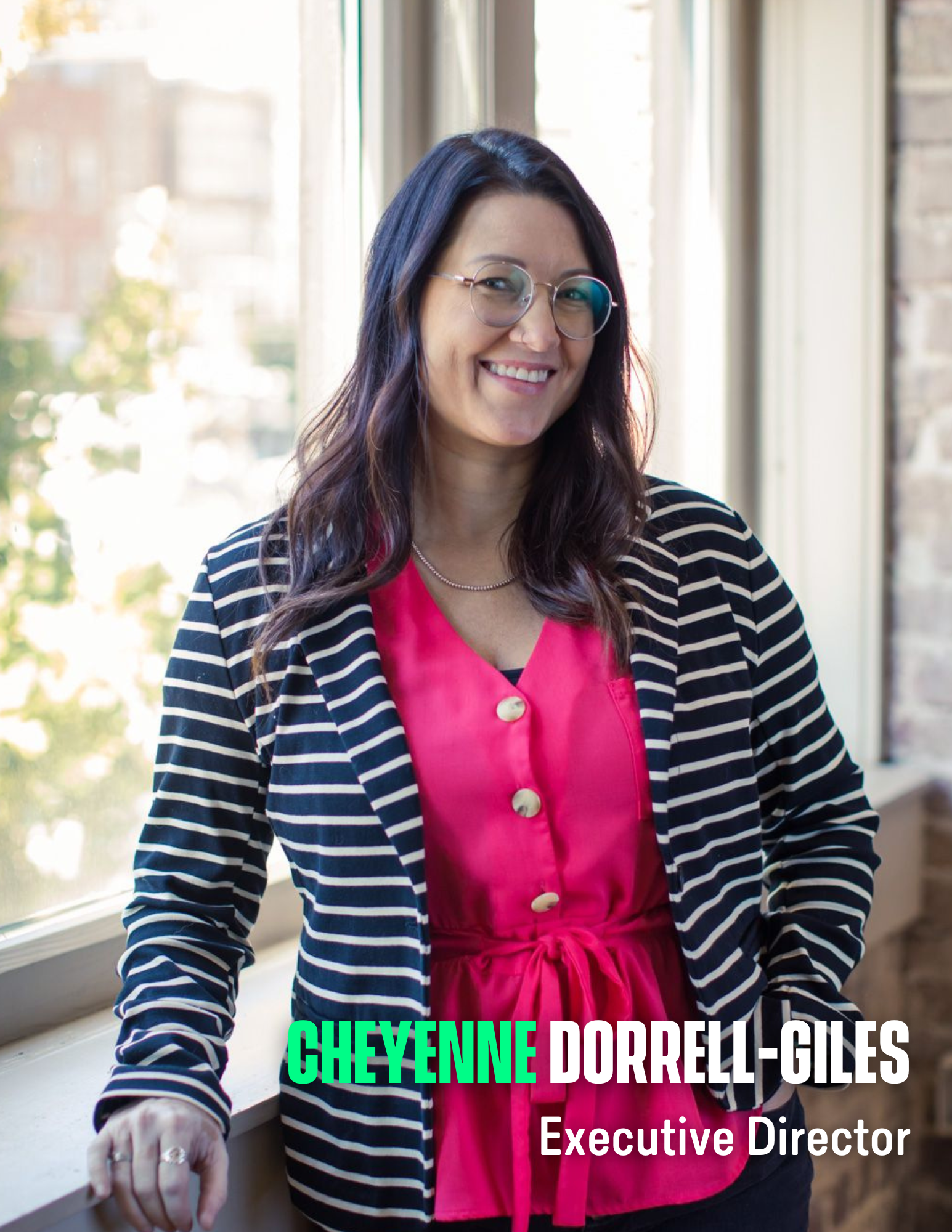 Photo of a woman with long dark hair, glasses and a pink blouse with striped blazer standing in front of a window.
