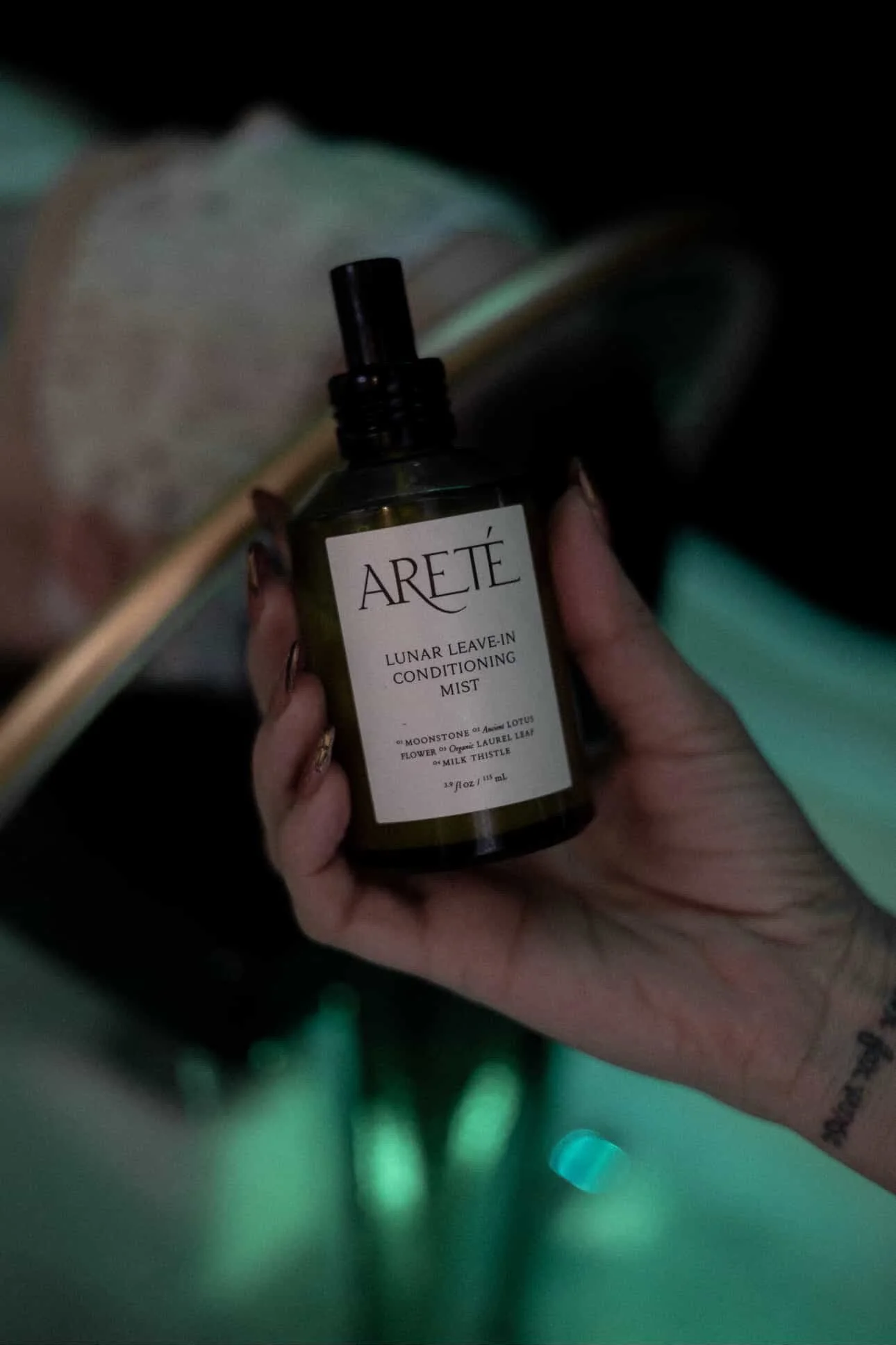 A hand holding a brown bottle of Areté Lunar Leave-in Conditioning Mist with a black spray nozzle, with an open book in the background and out-of-focus green and blue lighting.