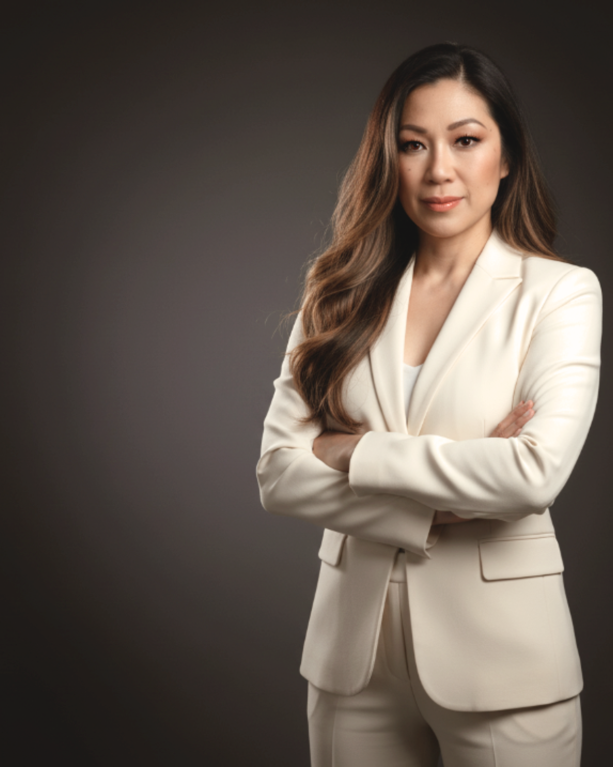 A woman with long, wavy brown hair wearing a cream-colored blazer and pants, standing with arms crossed against a dark gray background.