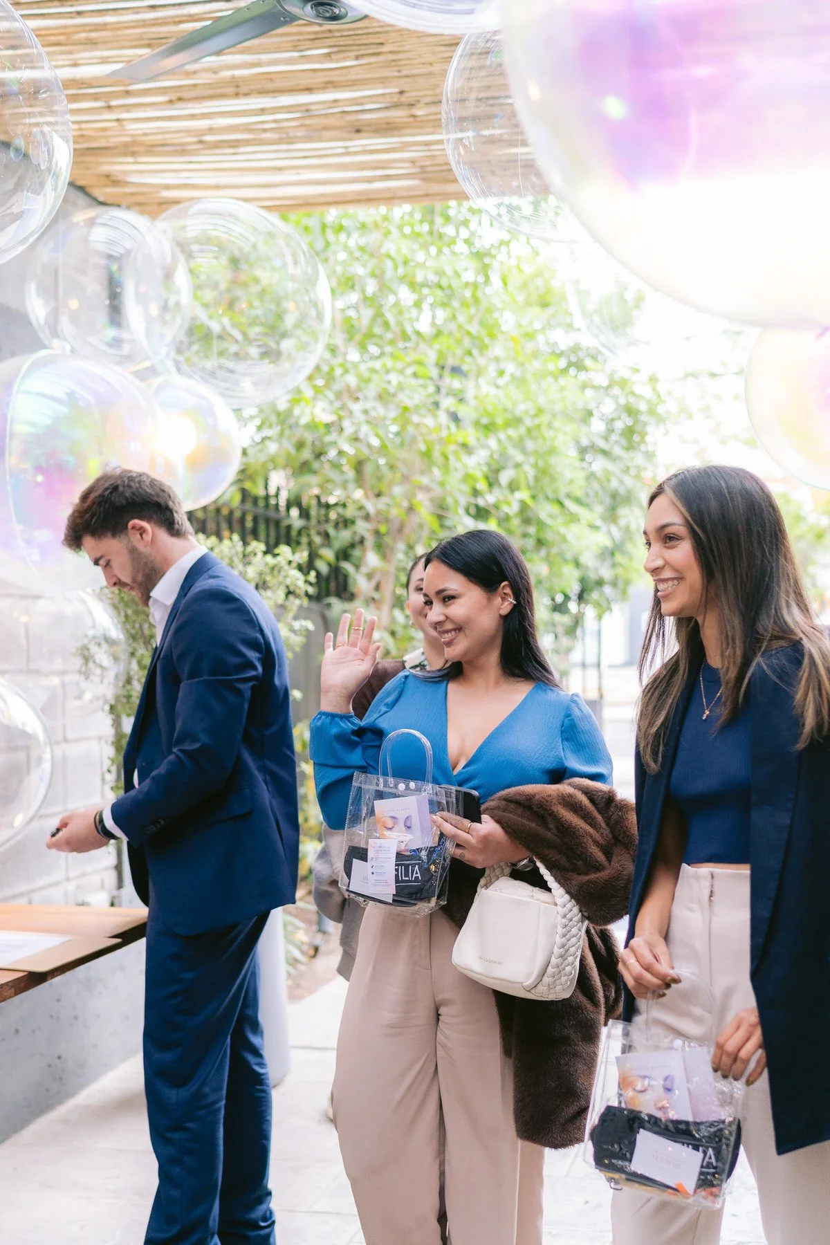 Grupo de personas sonrientes en un evento exterior decorado con globos transparentes iridiscentes, algunas sosteniéndose bolsas con souvenirs y otros interactuando.
