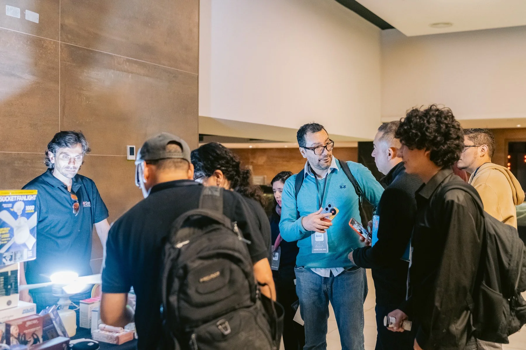 Grupo de personas conversando en un evento, algunos con mochilas y usando gafas, en un interior moderno con paredes de color marrón.