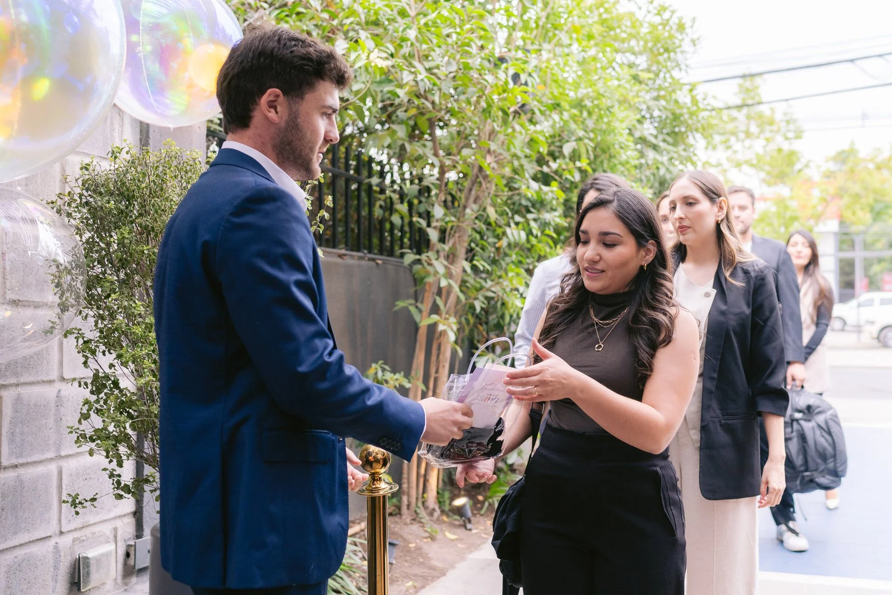 Una mujer recibe una bolsa con regalos de un hombre vestido con traje azul. La escena parece ser en un evento o celebración, con varias personas en línea y decoraciones de globos en el fondo.