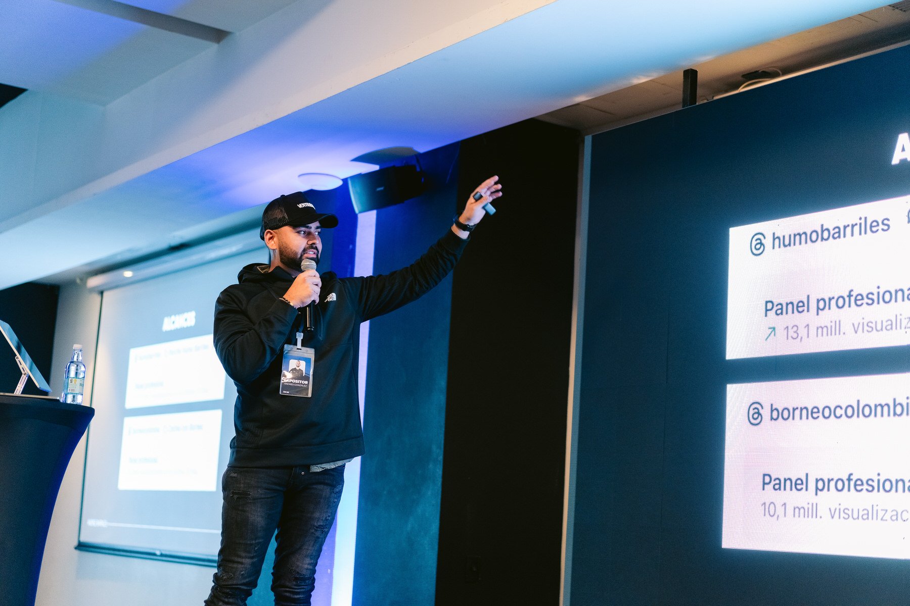 Hombre con gorra y chaqueta negra dando una presentación frente a una pantalla en un salón de conferencias.