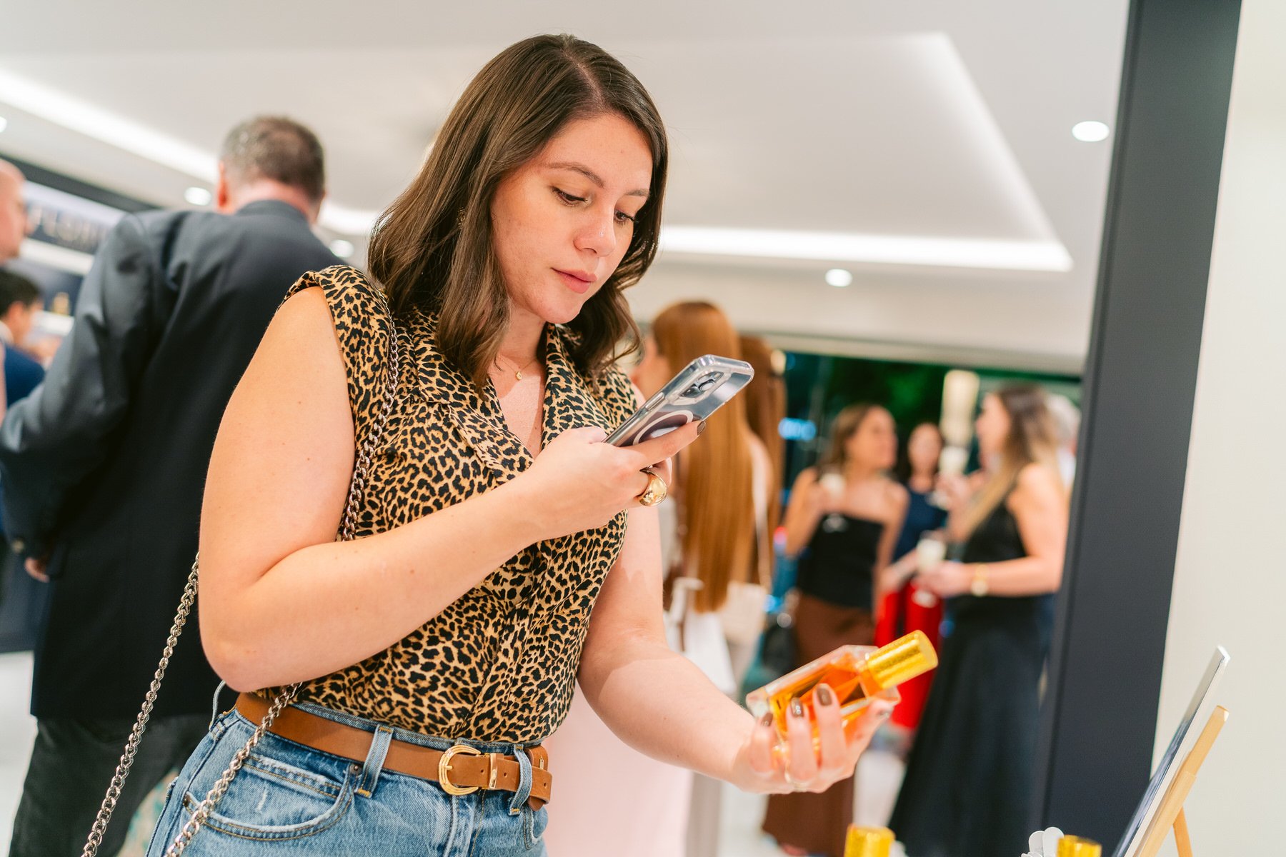 Joven mujer con blusa de animal print y jeans, usando su teléfono. En el fondo, varias personas conversando en un lugar público.
