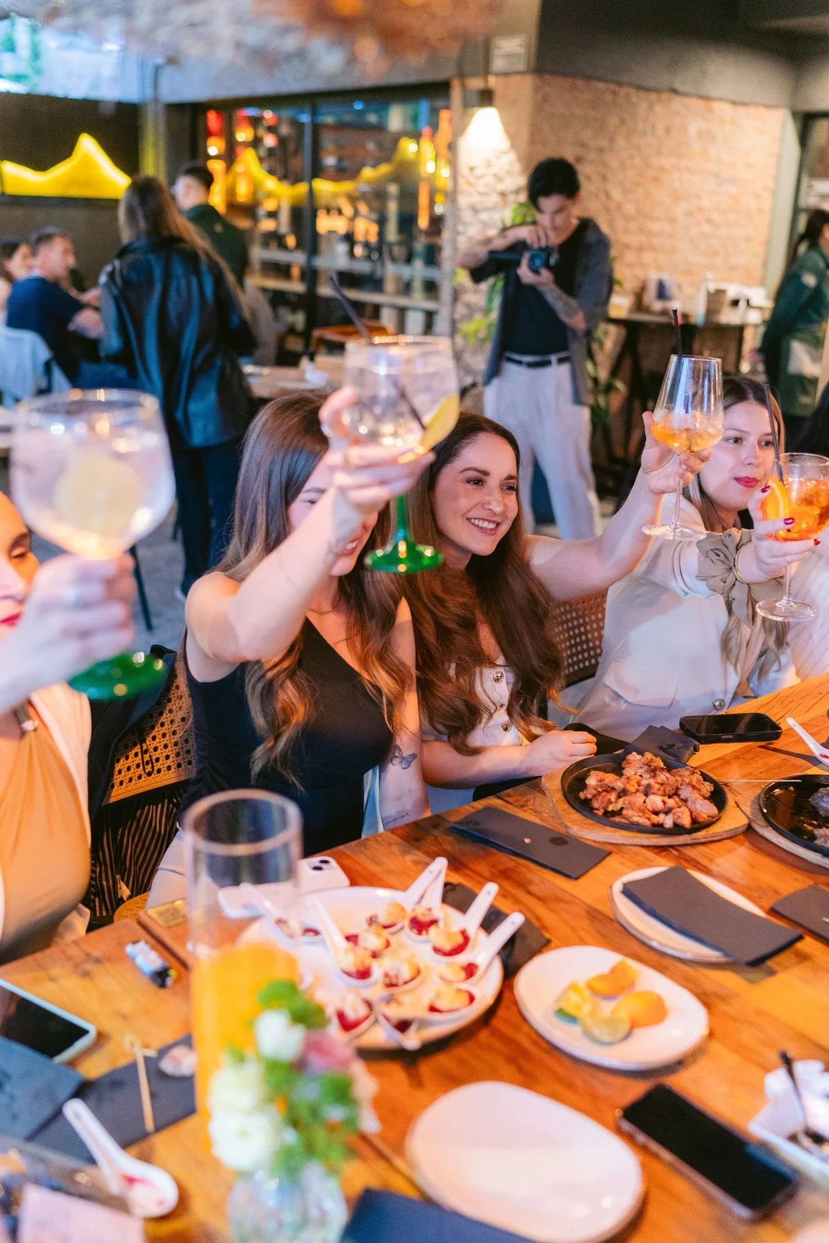Grupo de personas en una reunión en un restaurante, levantando copas con bebidas, con comida en la mesa y ambiente escolar.