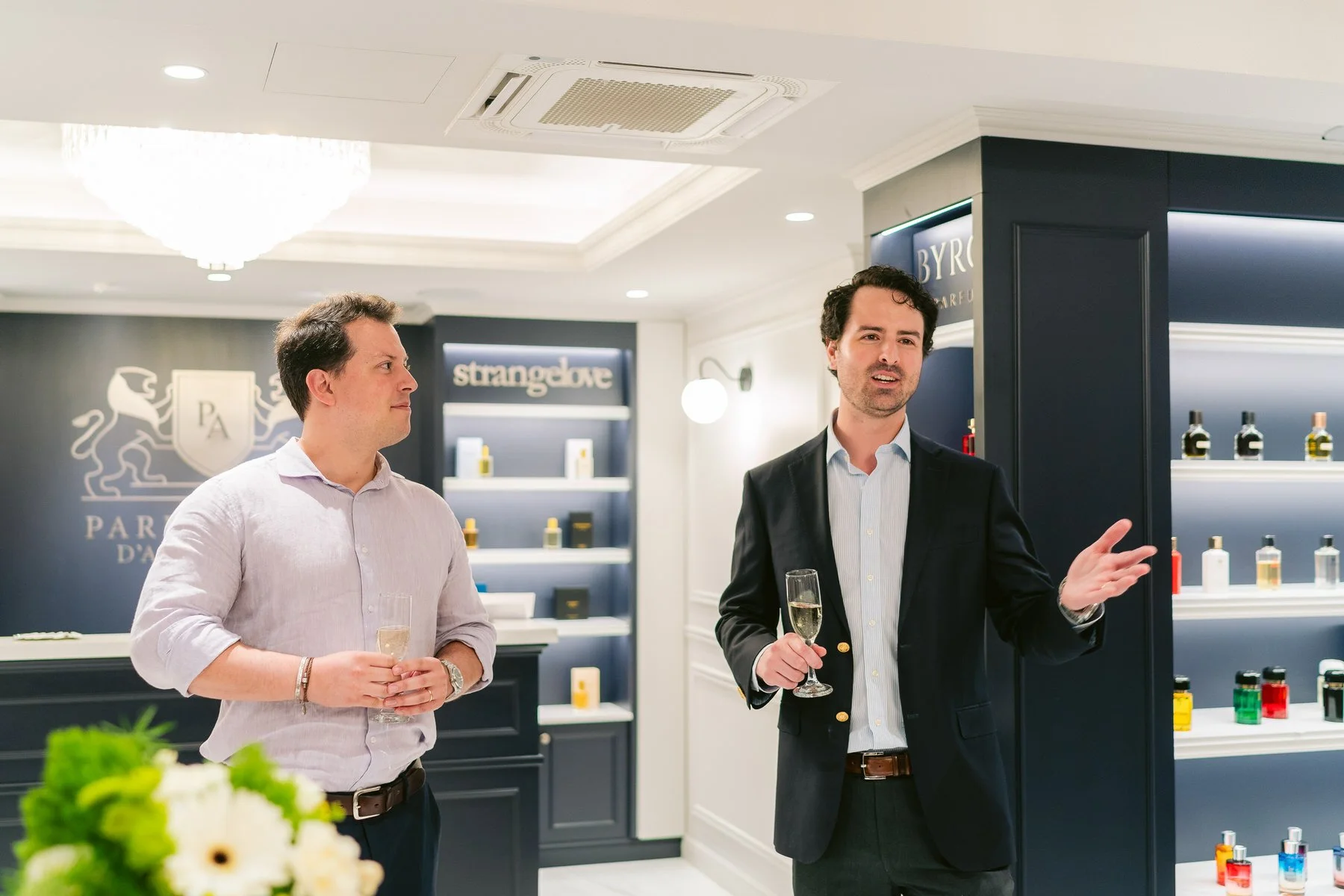 Dos hombres en una tienda o sala de exhibiciones, uno habla y el otro escucha. Ambos sostienen copas con bebidas espumosas. La tienda tiene estanterías con frascos y botellas, y un logotipo en la pared con un escudo, una figura de león y las palabras