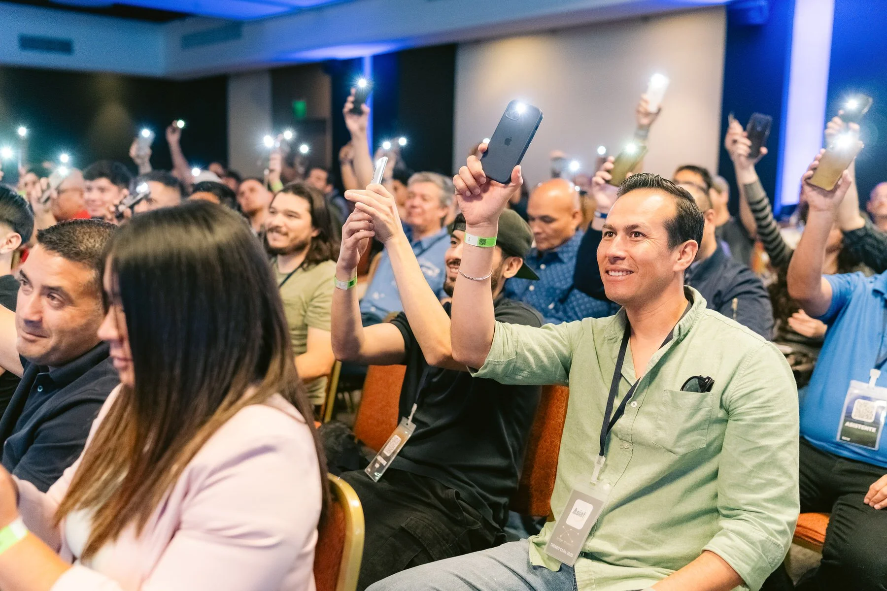 Hombre en la audiencia levantando un teléfono móvil para grabar o tomar fotos en un evento con muchas personas, muchas con teléfonos encendidos y con velas encendidas.