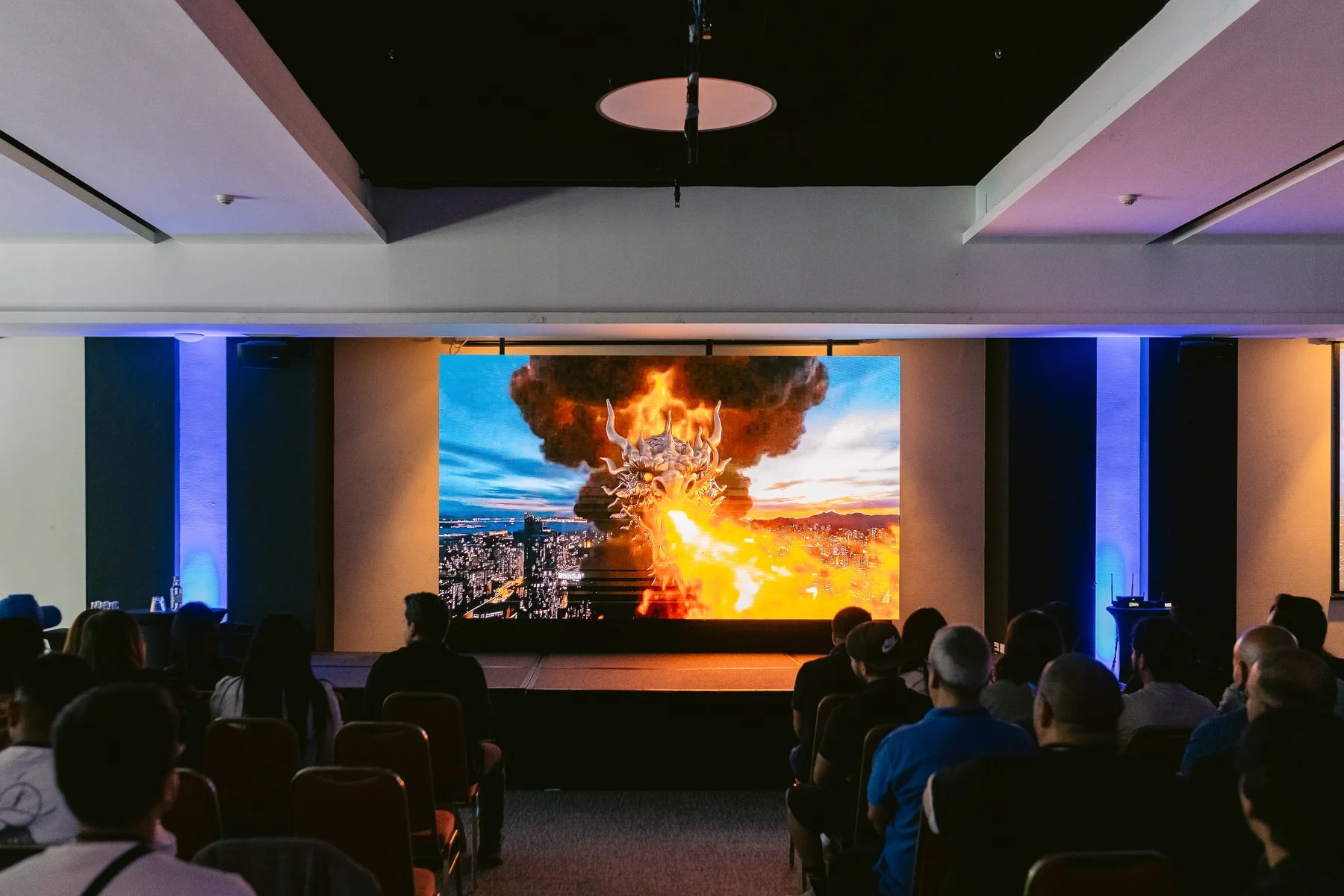 Audiencia viendo una pantalla grande que muestra una escena de un dragón lanza fuego en una ciudad con una explosión en el fondo.