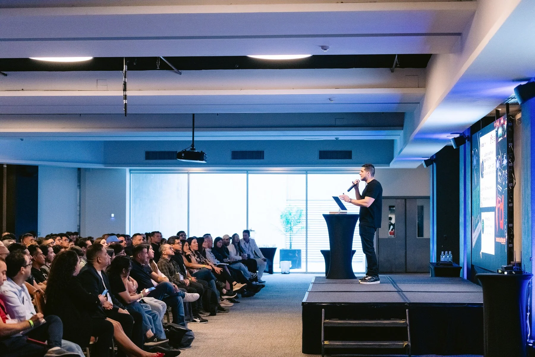 Un orador en un escenario dando una presentación frente a una audiencia atentos en un salón amplio con luz natural.
