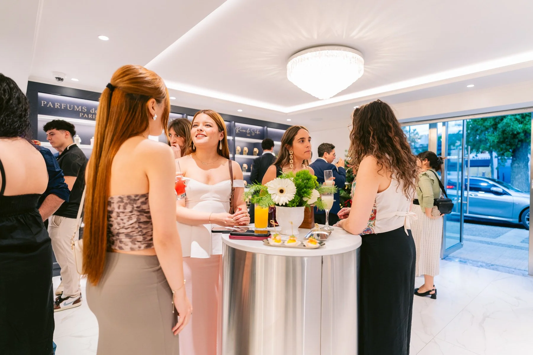 Grupo de mujeres conversando alrededor de una mesa en una fiesta o evento social dentro de un lugar elegante, con decoraciones y bebidas en mano.