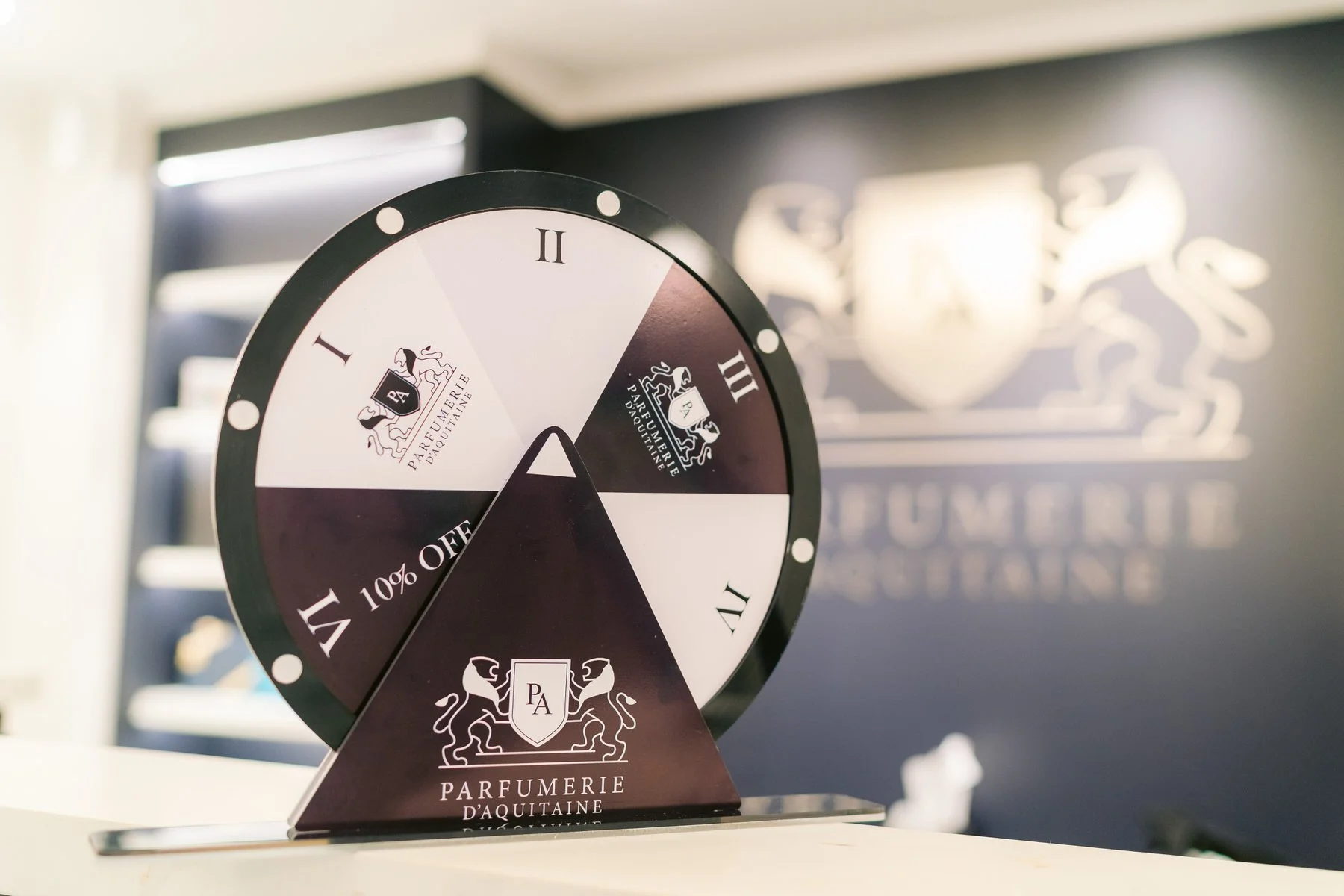 Reloj de rueda con segmentos blancos, negros y morados, con logotipo y texto de 'Parfumerie d'Aquitaine', en fondo de pared con logotipo y escudo en colores claros.