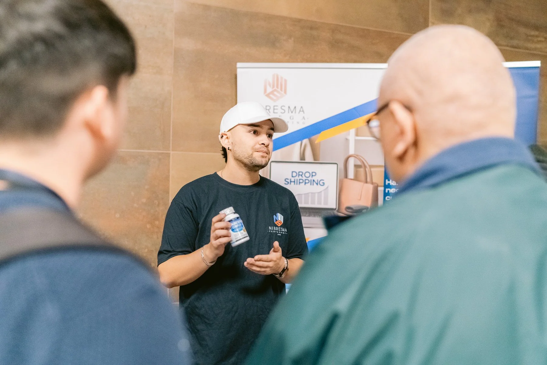 Un joven hablando con dos adultos en una conferencia, sosteniendo una botella de medicamento y frente a un stand con cartel que dice "Drop Shipping" y logo de una empresa llamada NIBRESMA.
