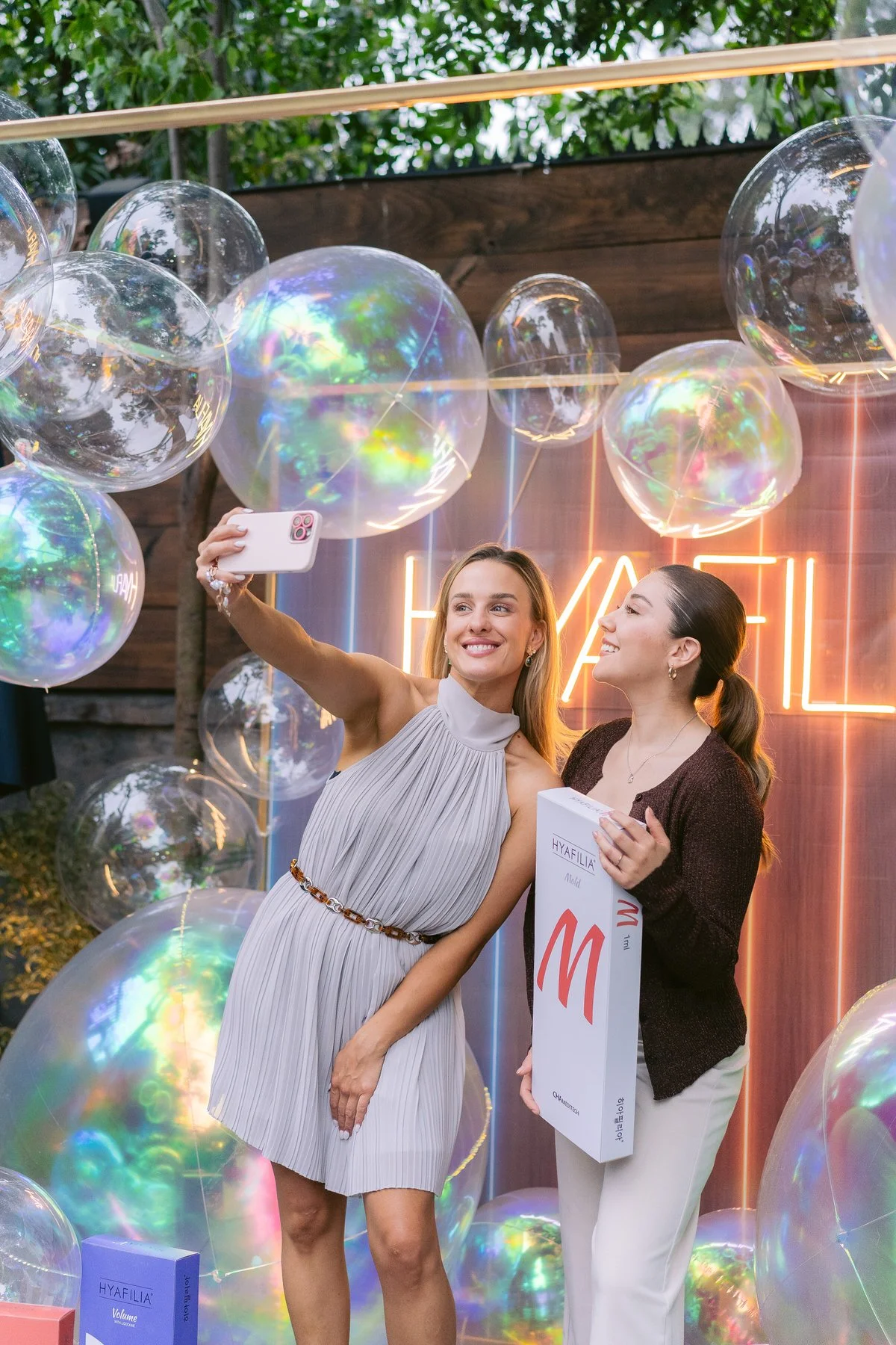 Dos mujeres tomando una selfie en una fiesta, rodeadas de globos traslúcidos y un fondo con neón que dice 'HYAFILIA'.