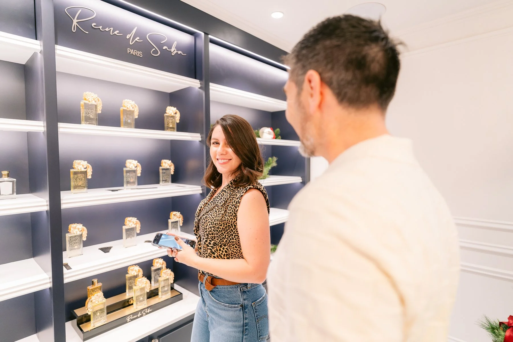Una mujer con blusa de animal print y jeans sosteniendo un teléfono, sonriendo frente a una repisa con frascos de perfume, en una tienda llamada Rive d' Sable en París.