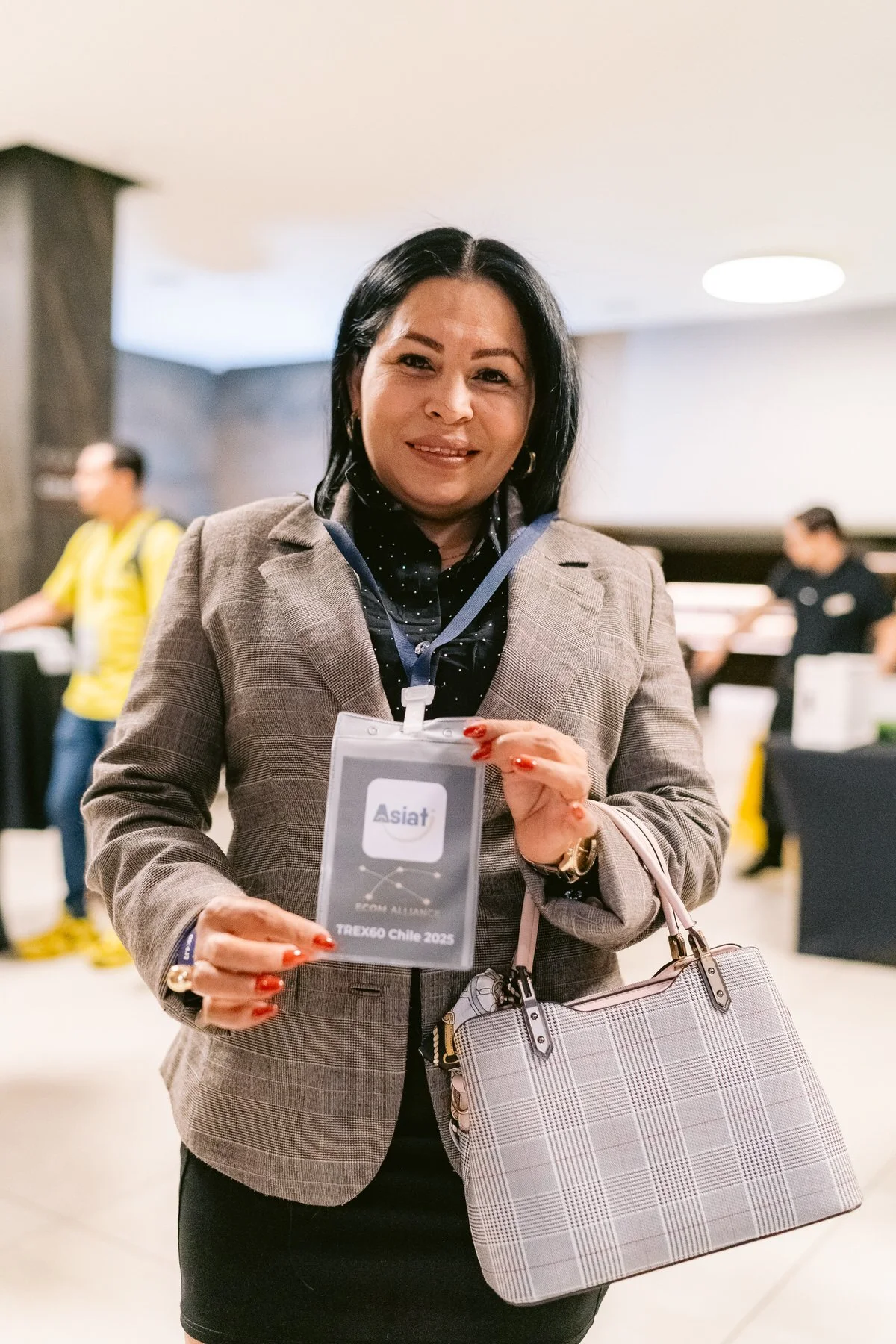 Una mujer sonriente con blazer de cuadros sostiene una acreditación de conferencia en una evento. Lleva bolso a cuadros y accesorios dorados, fondo de sala con personas.