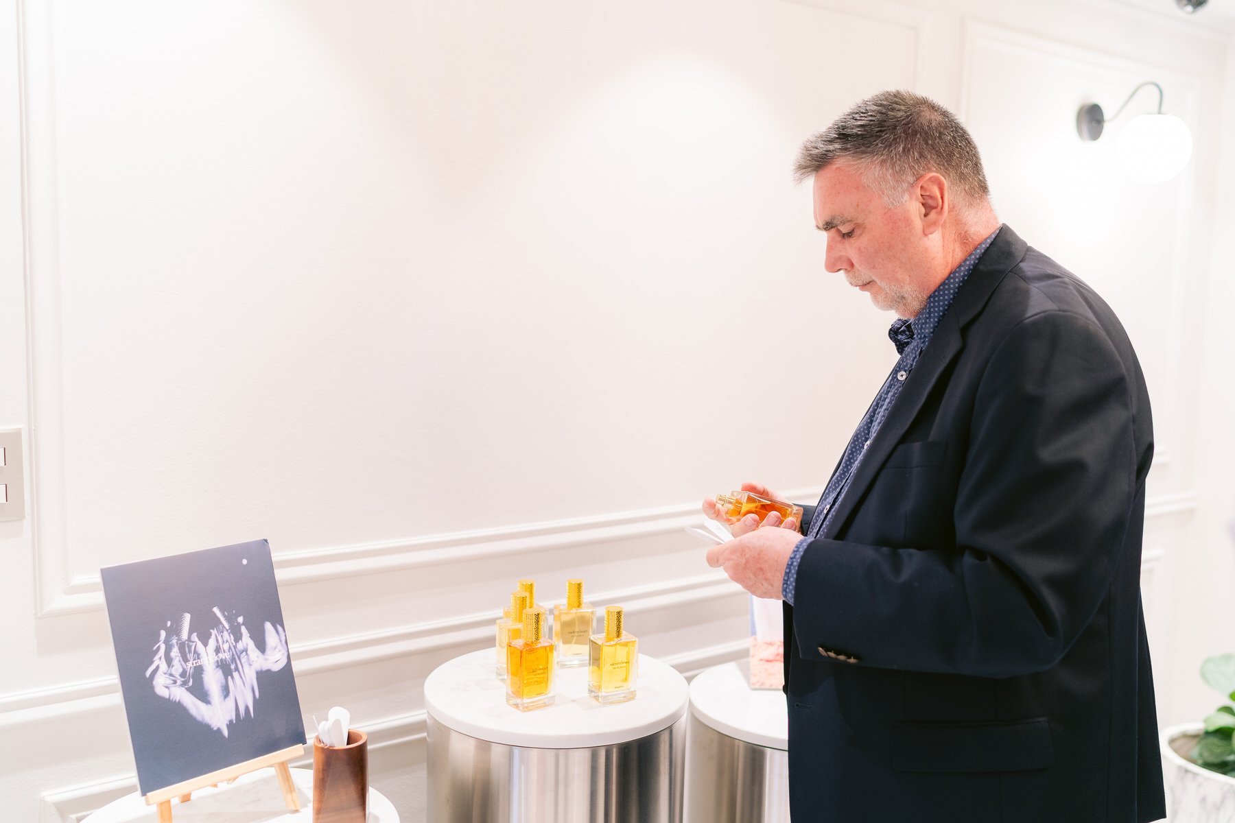 Hombre con barba y cabello canoso vestido con saco negro, observando frascos de perfume en una mesa redonda blanca en una tienda o exhibición de perfumes.